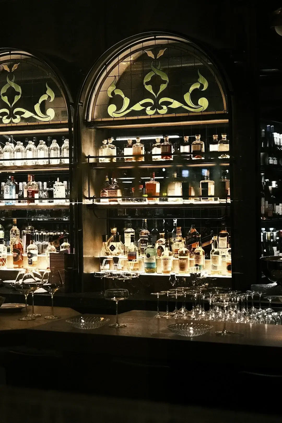 Backlit display of various bottles of liquor on shelves behind a bar counter, with empty wine glasses and cocktail glasses in the foreground.