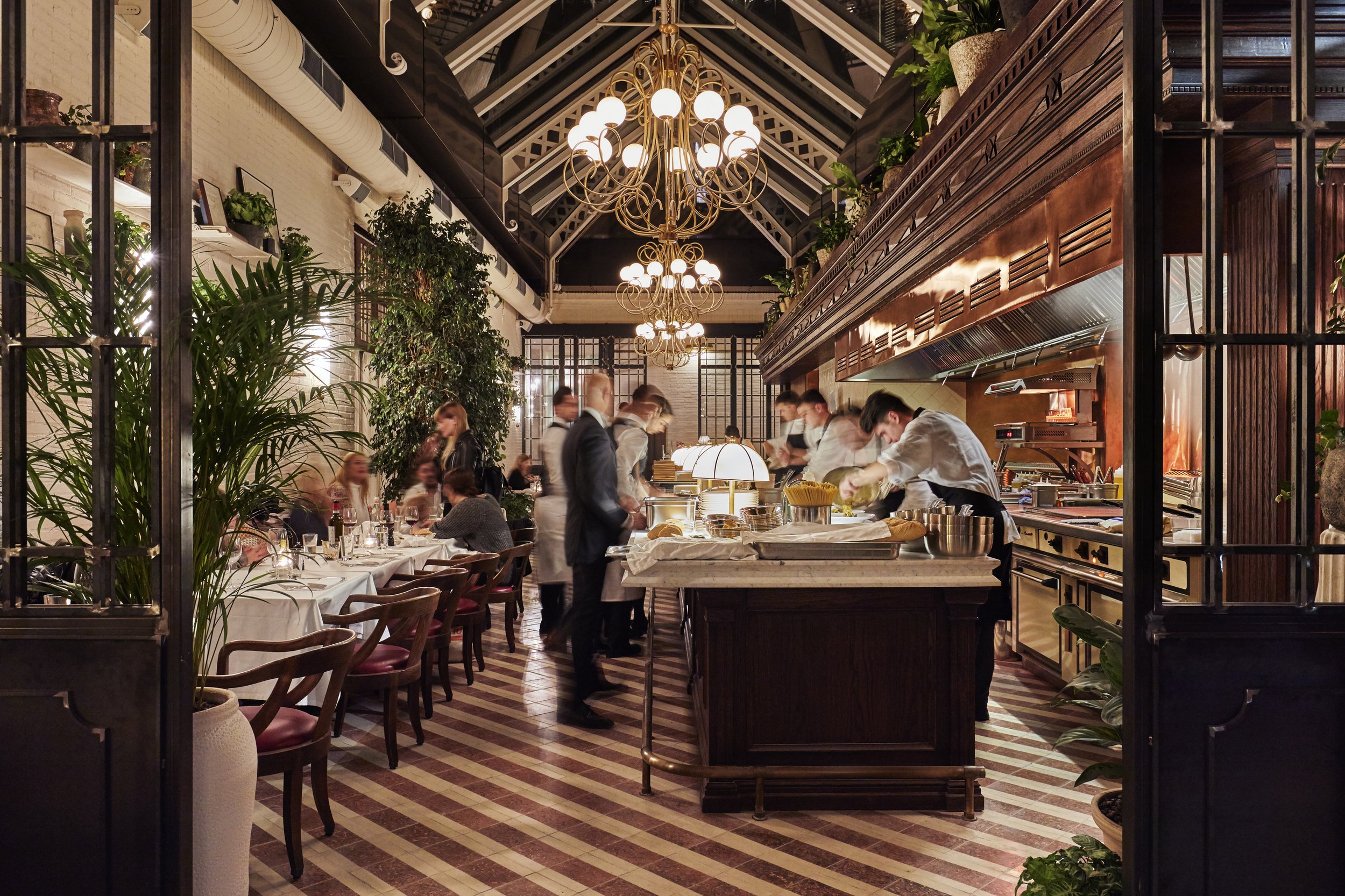 Elegant restaurant with dining table, people dining and waiters serving, decoration with plants and chandeliers.