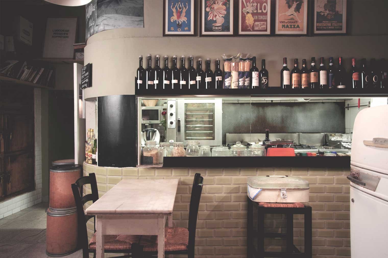 Interior of an open kitchen in a restaurant with wine bottles on a shelf, wooden table with chairs, and visible cooking utensils. Retro decor with posters on the wall.