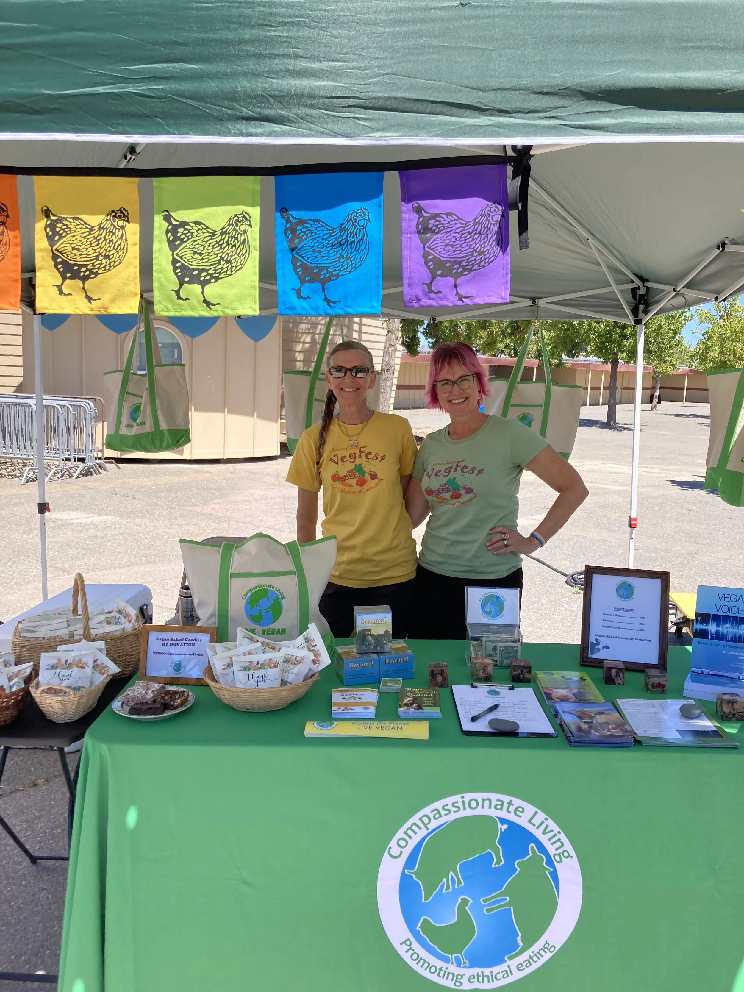 Two women standing behind a table at an outdoor vegan event, with colorful banners and informational materials promoting ethical living and veganism.