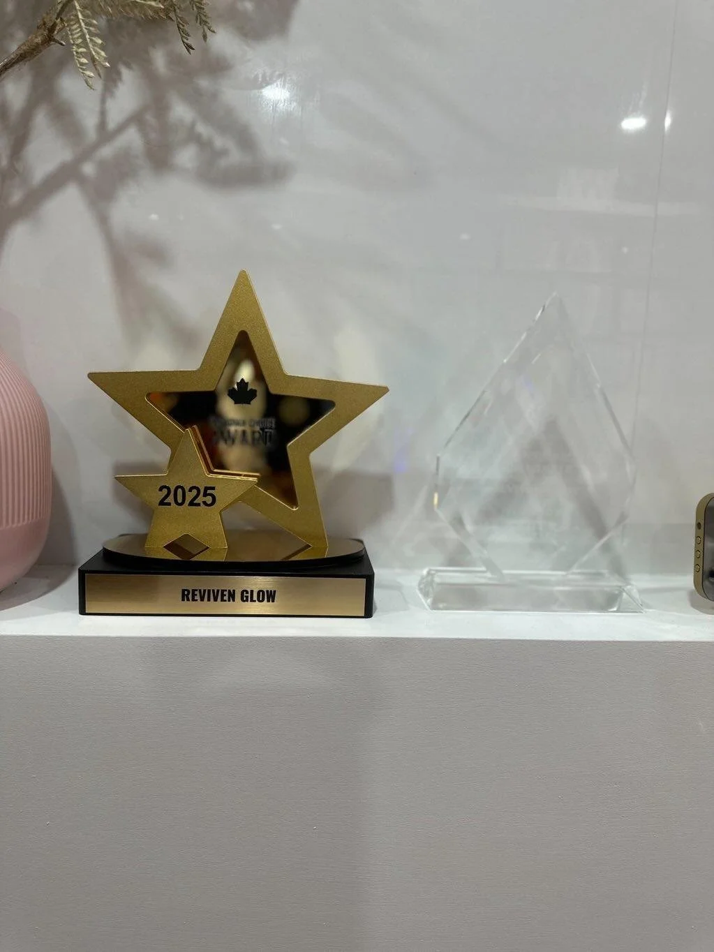 A gold star trophy labeled 'Reviven Glow' with the year 2025 on a black base, placed on a white shelf next to a glass award, against a white background.