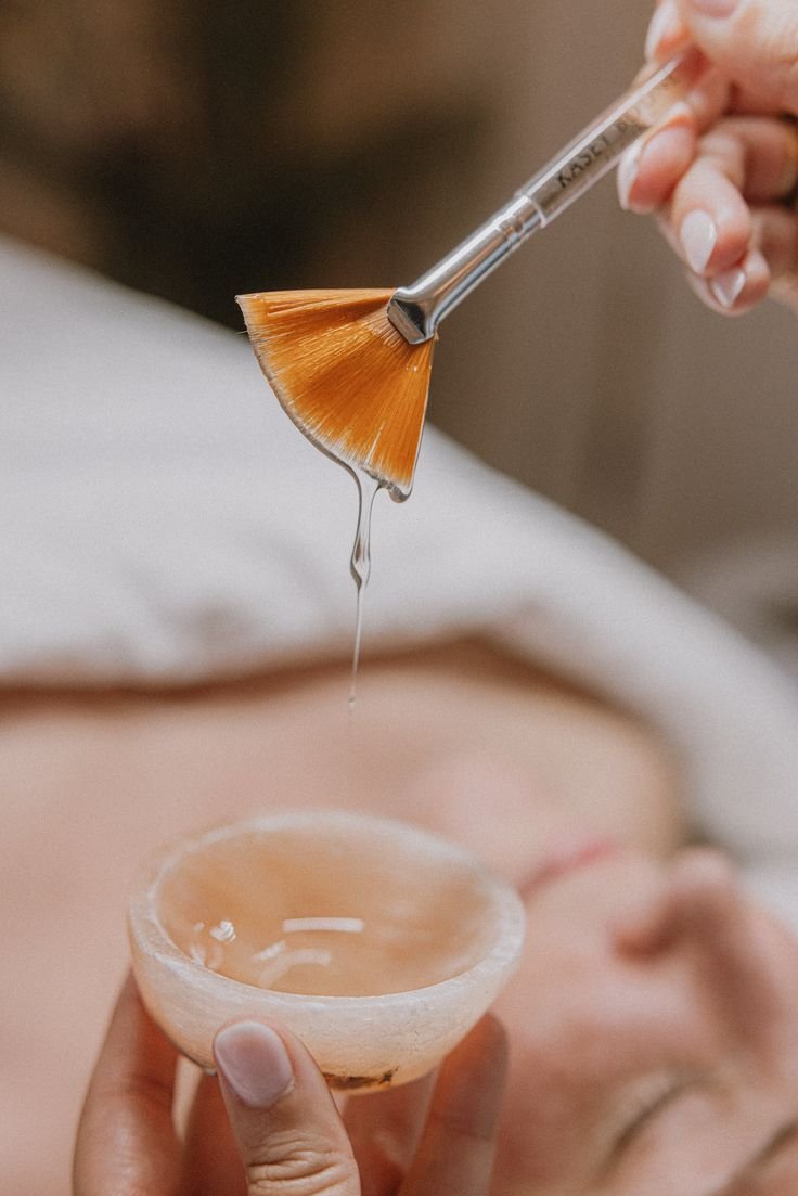 A person holding a small cup with a wax fan brush applying wax, with a drop of liquid wax hanging from the brush.