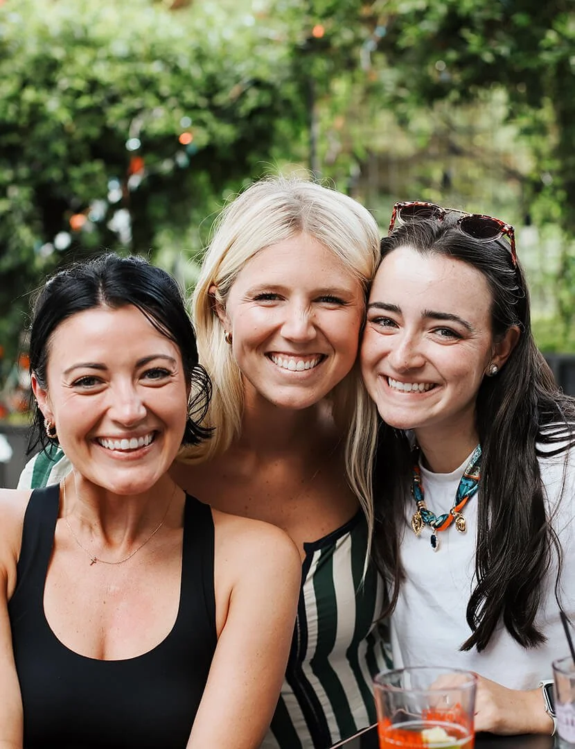 Three women smiling outdoors in a garden, with trees and string lights in the background.
