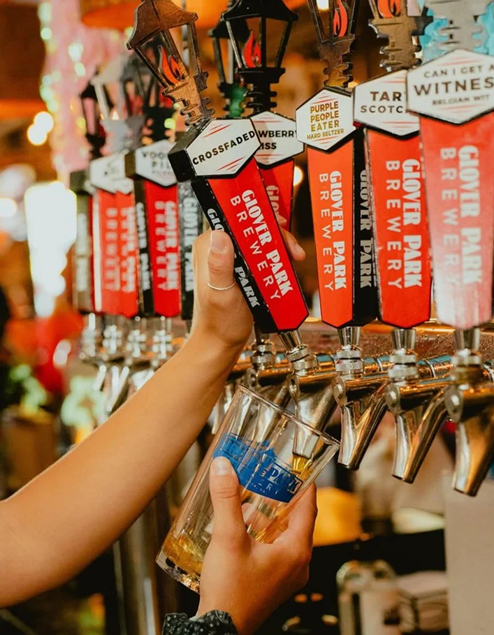 A person filling a glass with beer from a tap at a bar or brewery, with multiple beer taps labeled with different flavors.