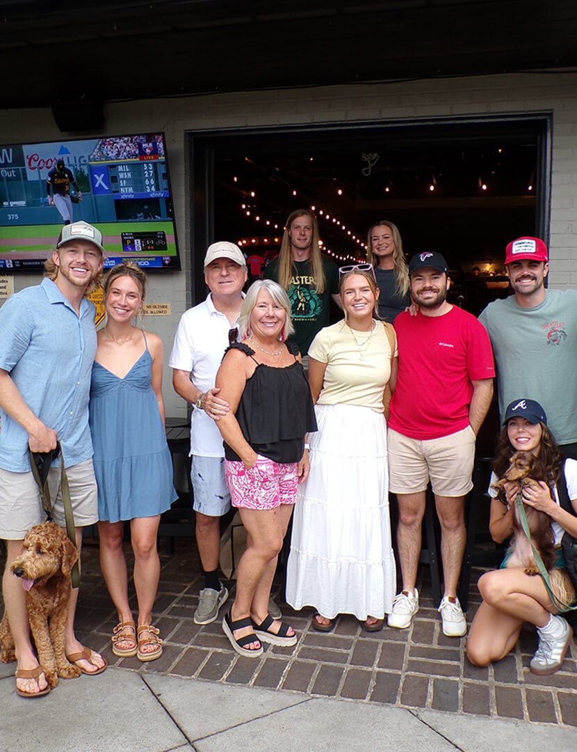Group of people posing outside a restaurant or bar with a TV screen showing a baseball game in the background.