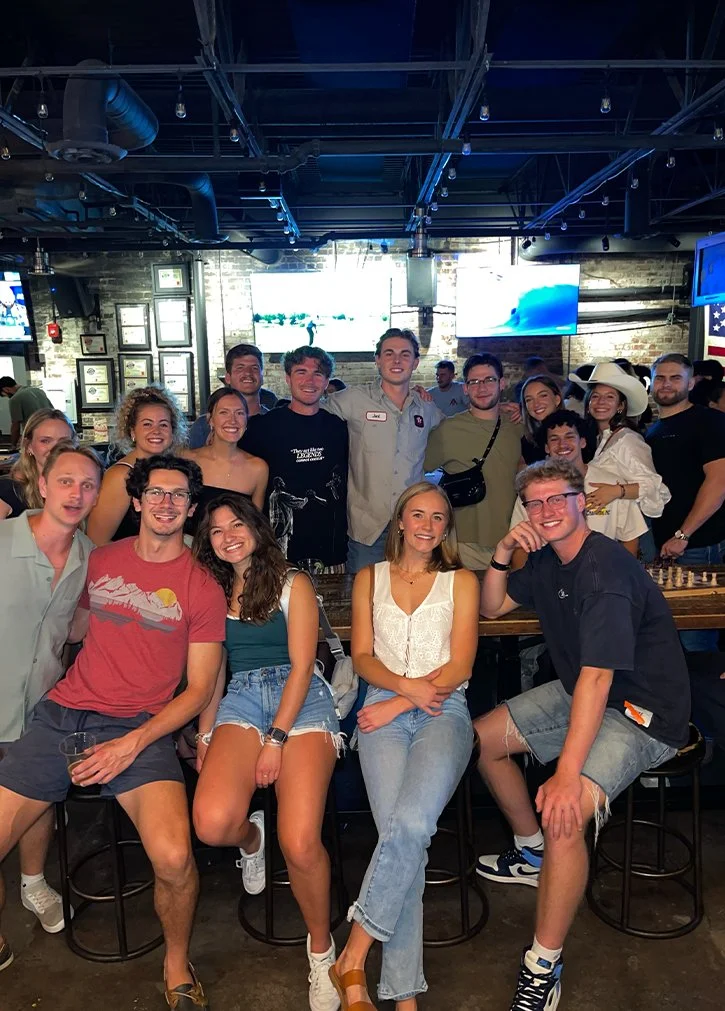 A group of young adults smiling and posing for a photo in a bar or restaurant setting with televisions and framed pictures on the wall in the background.