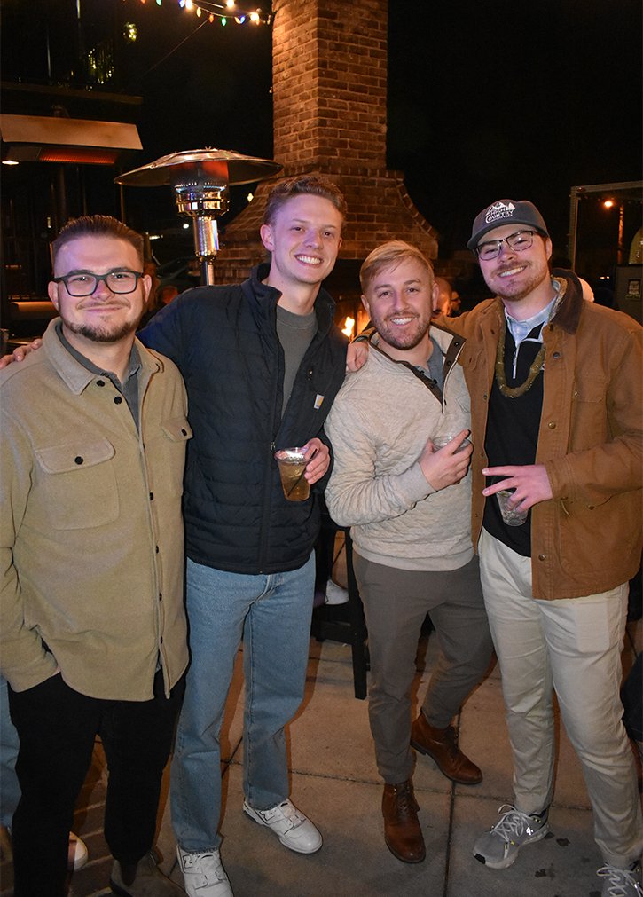 Four men standing together outdoors at night, smiling, holding drinks, near a brick chimney and outdoor heater, with string lights overhead.