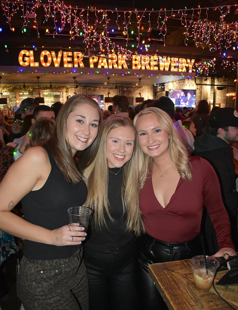 Three women smiling and posing at Glover Park Brewery with colorful string lights and a crowded bar in the background.