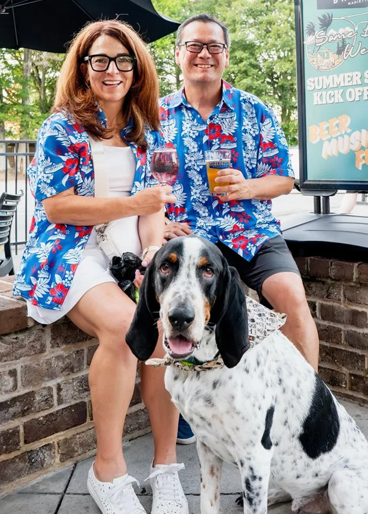 Two smiling adults, a woman with glasses and a man, sitting outside at a bar or restaurant, holding drinks, with a large black and white dog sitting in front of them. The woman is wearing a white dress with a blue Hawaiian shirt, and the man is weari