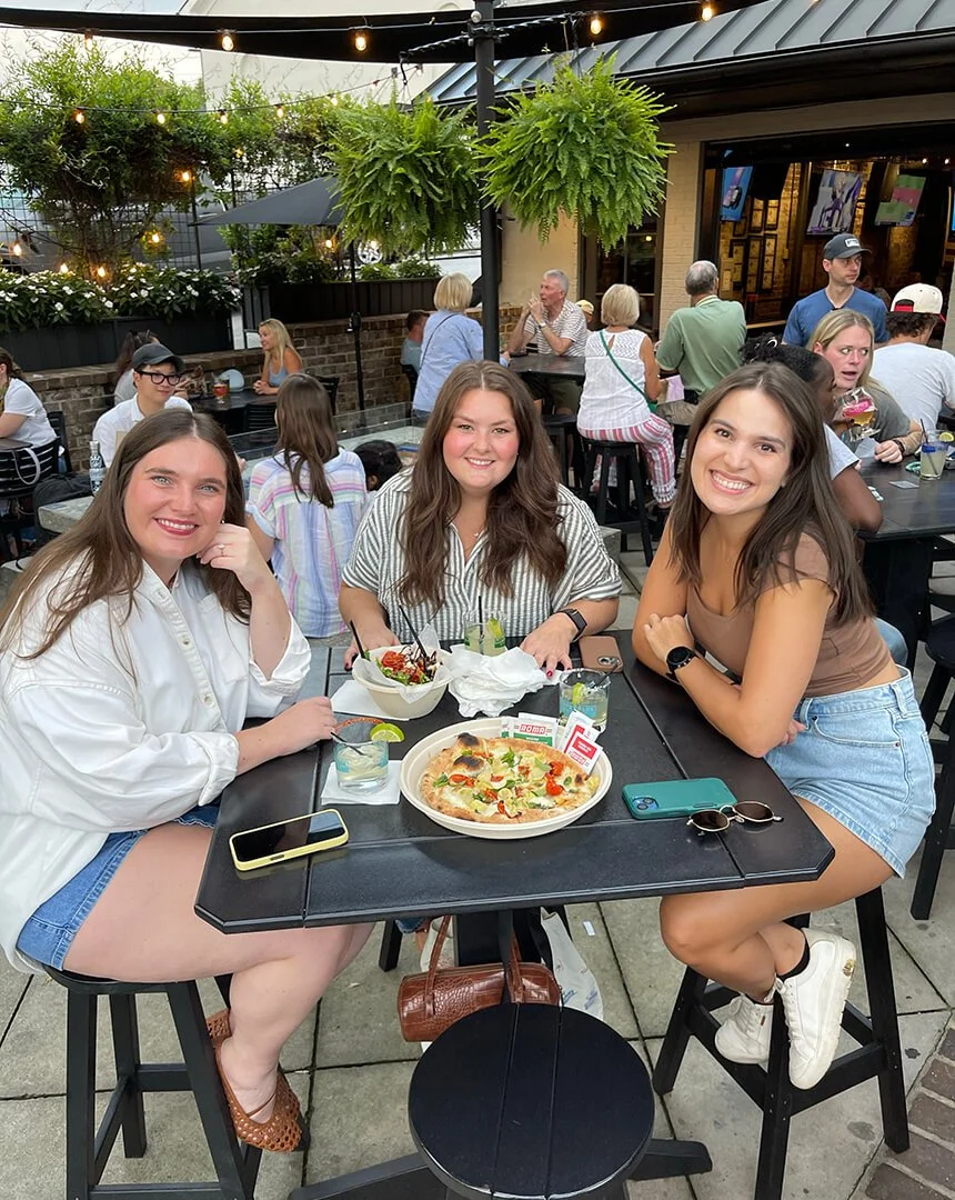 Three young women enjoying a meal and drinks at an outdoor restaurant or patio, with others dining in the background, some standing and talking, and lush greenery overhead.