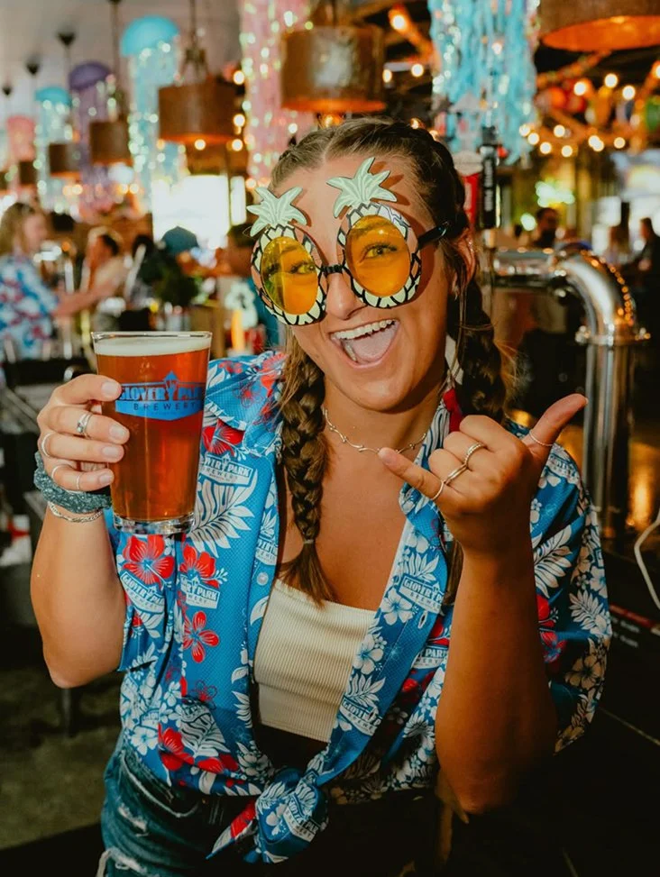 A woman wearing tropical-themed sunglasses and a colorful floral shirt, holding a glass of beer, smiling and making a shaka sign in a lively bar or pub with festive decorations and other patrons in the background.