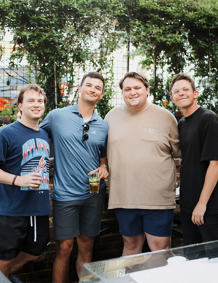 Four young men standing outdoors in front of green foliage, smiling and posing for a photo. One is holding a drink, and they are dressed casually.