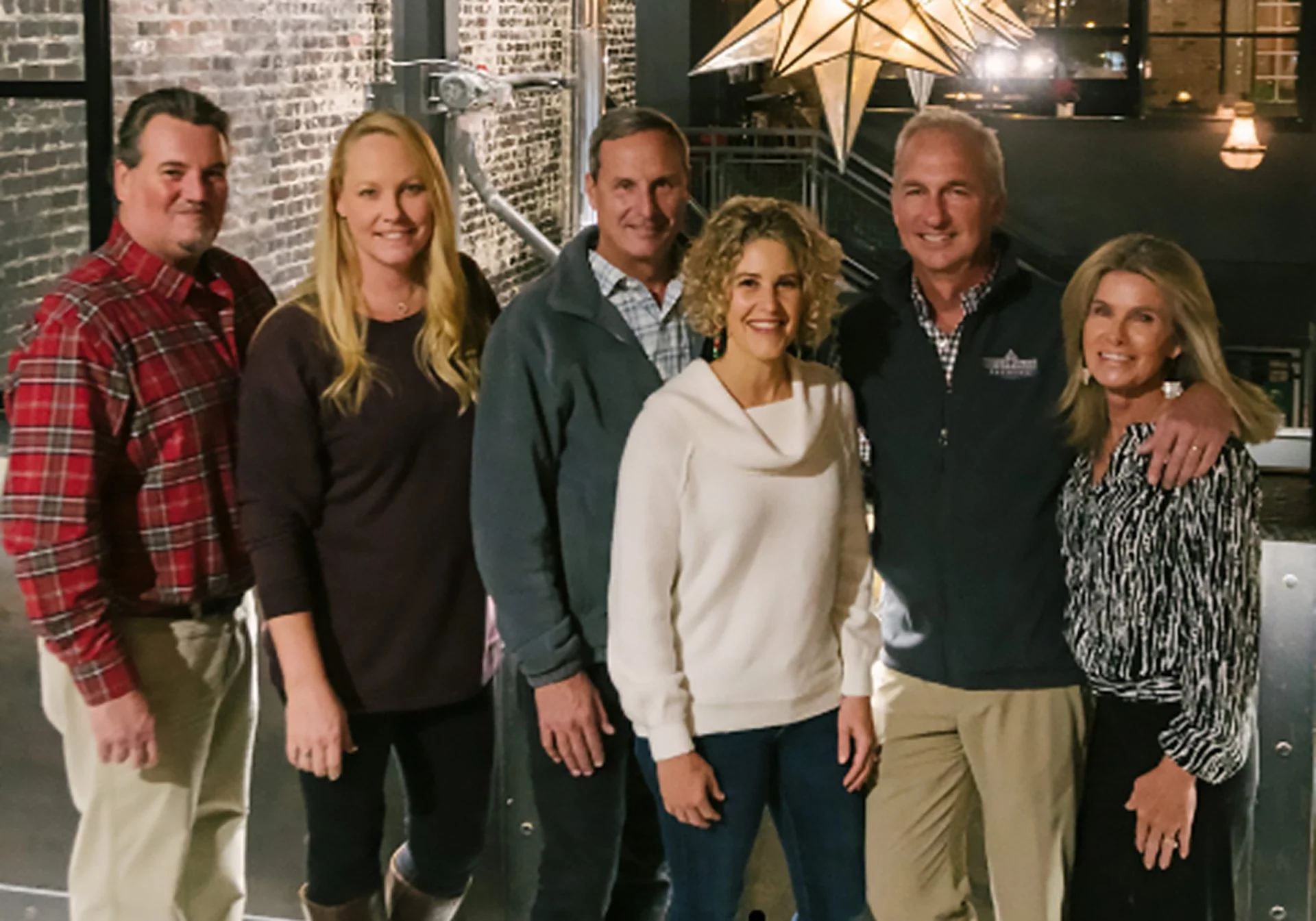 Group of six people posing indoors, with brick wall and decorative star light in background, smiling at camera.