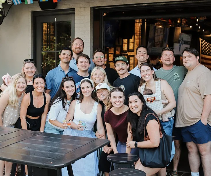 Group of 16 people smiling and posing for a photo outside a building, some holding drinks, during a social gathering or celebration.