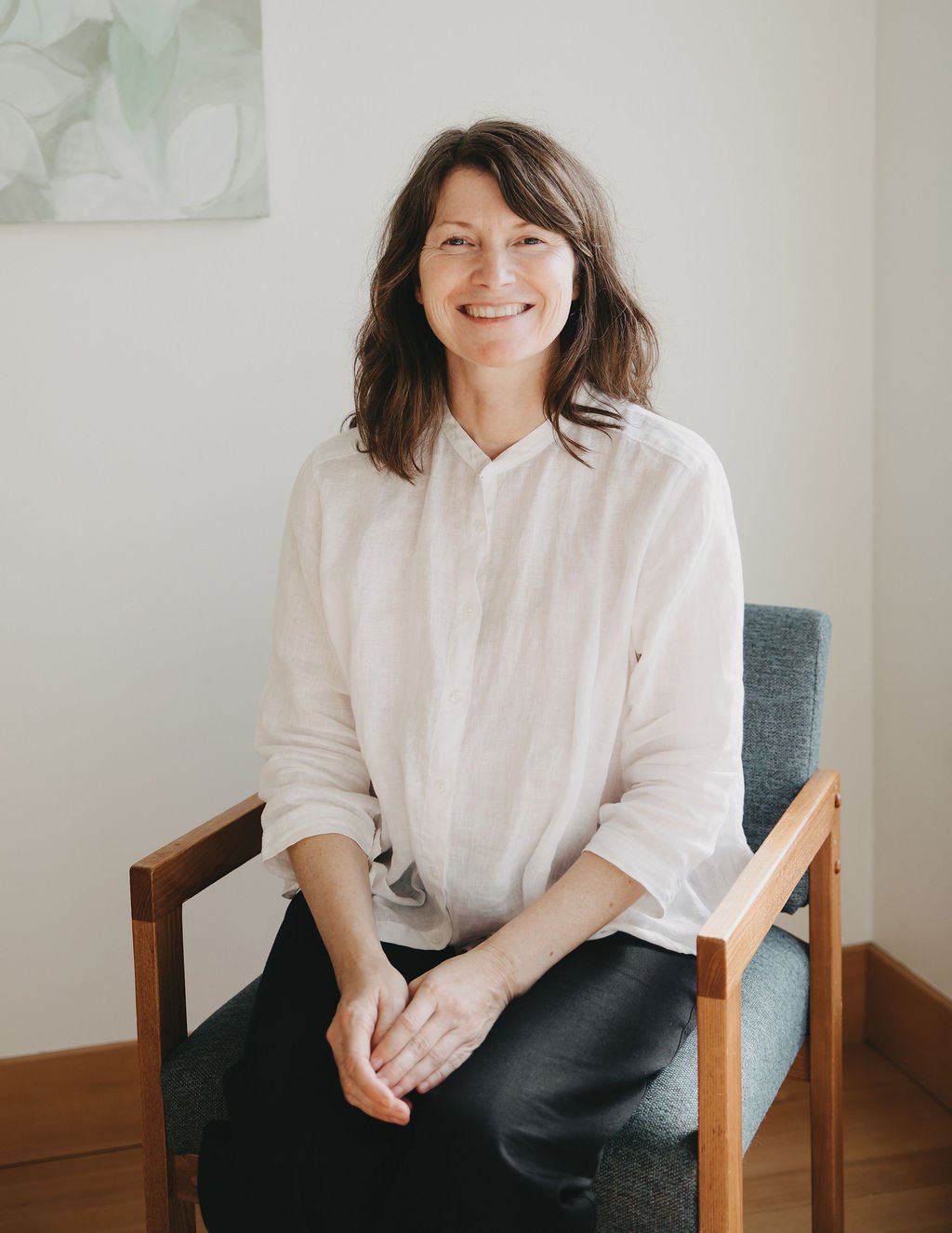 A woman with shoulder-length dark brown hair smiling and sitting on a wooden chair with a gray cushion, wearing a white shirt and black pants, in a brightly lit room with a light-colored wall and a painting in the background.