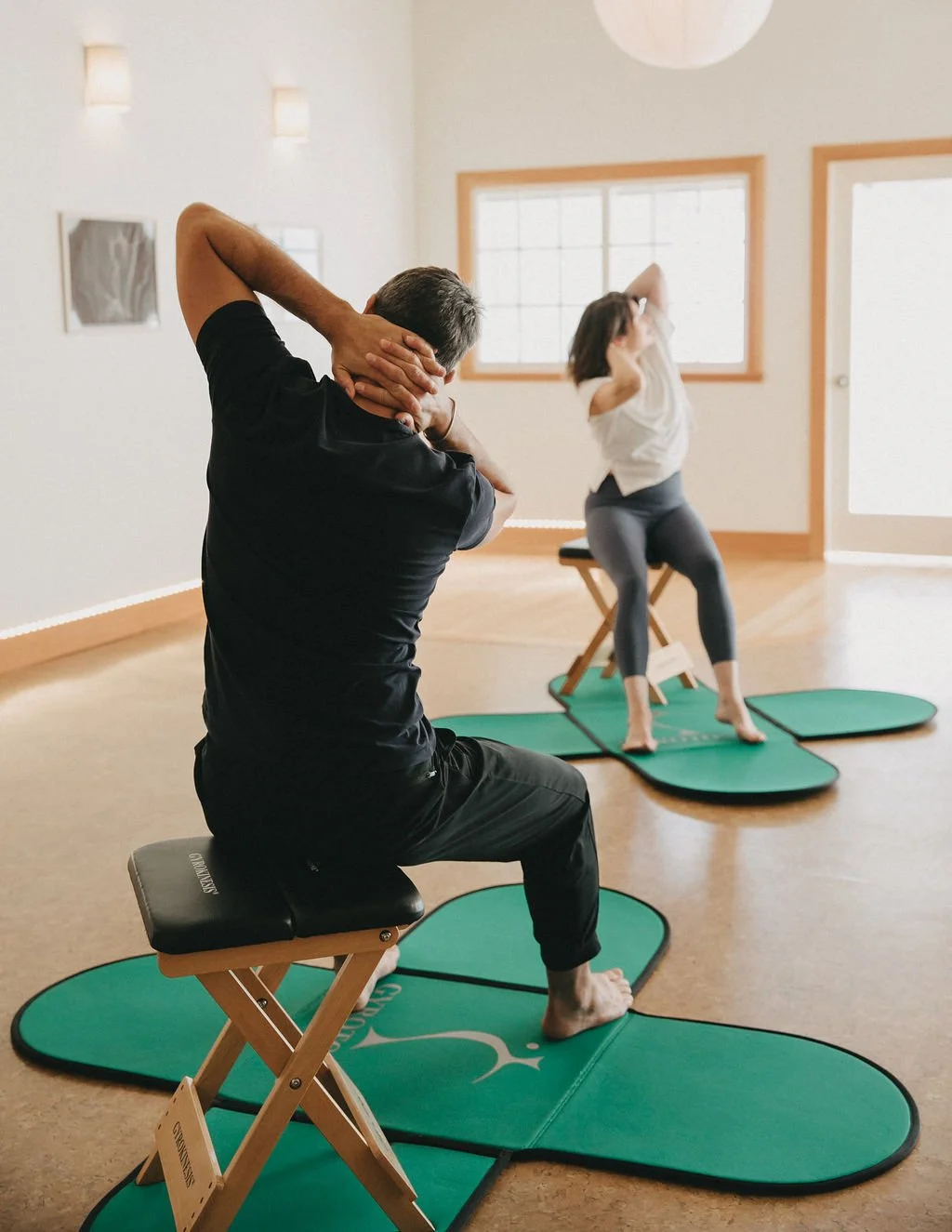 A group of women participating in a seated yoga class, following an instructor in a bright studio with wooden floors and large windows.