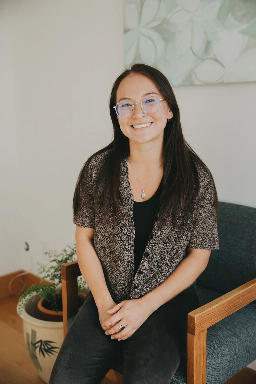 A young woman with long dark hair, glasses, and hoop earrings sitting on a wooden chair with gray cushions, smiling at the camera, with a potted plant and a white wall with green leaf artwork in the background.