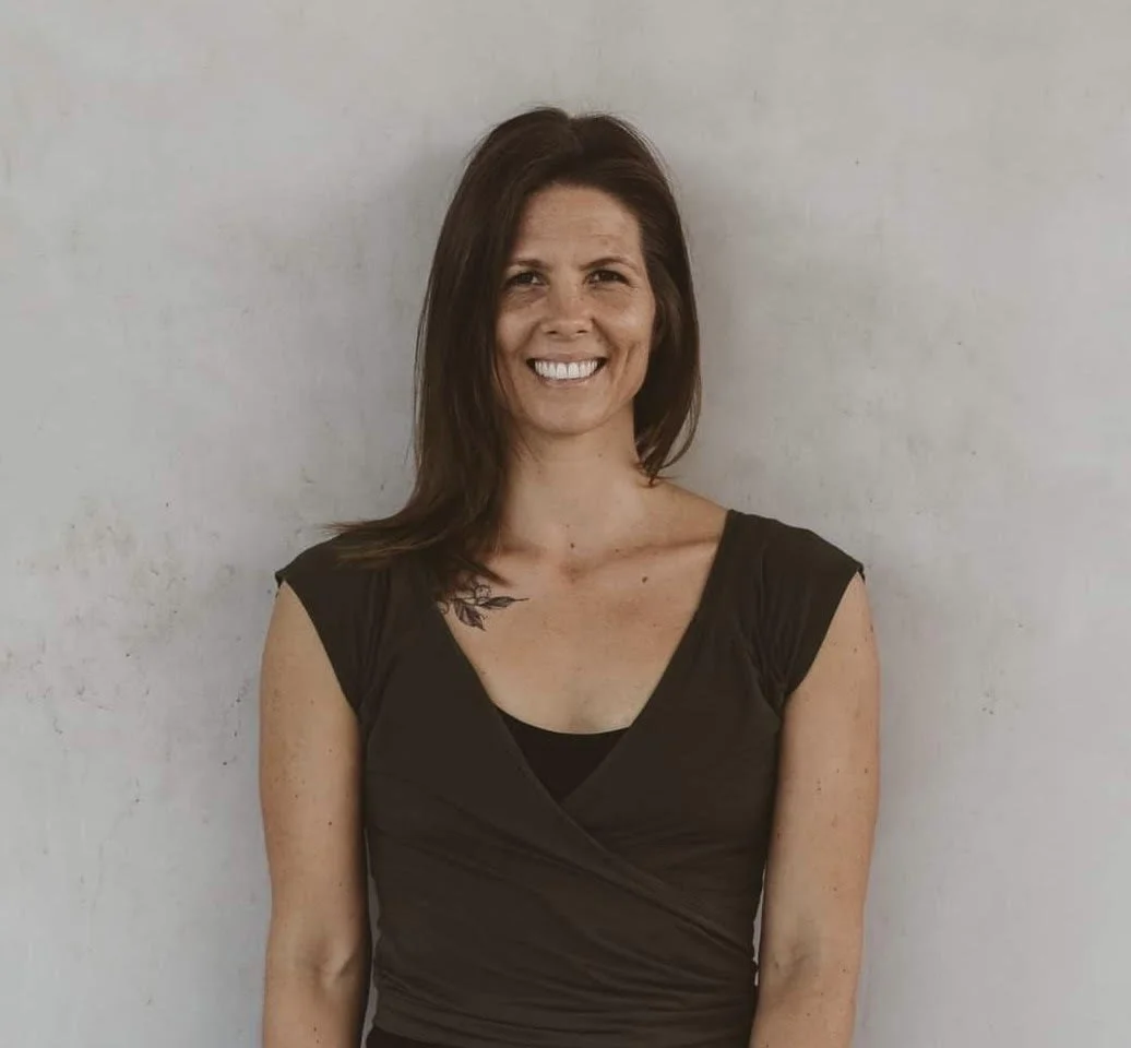 A woman with shoulder-length brown hair smiling, wearing a black top, standing against a plain light-colored wall.