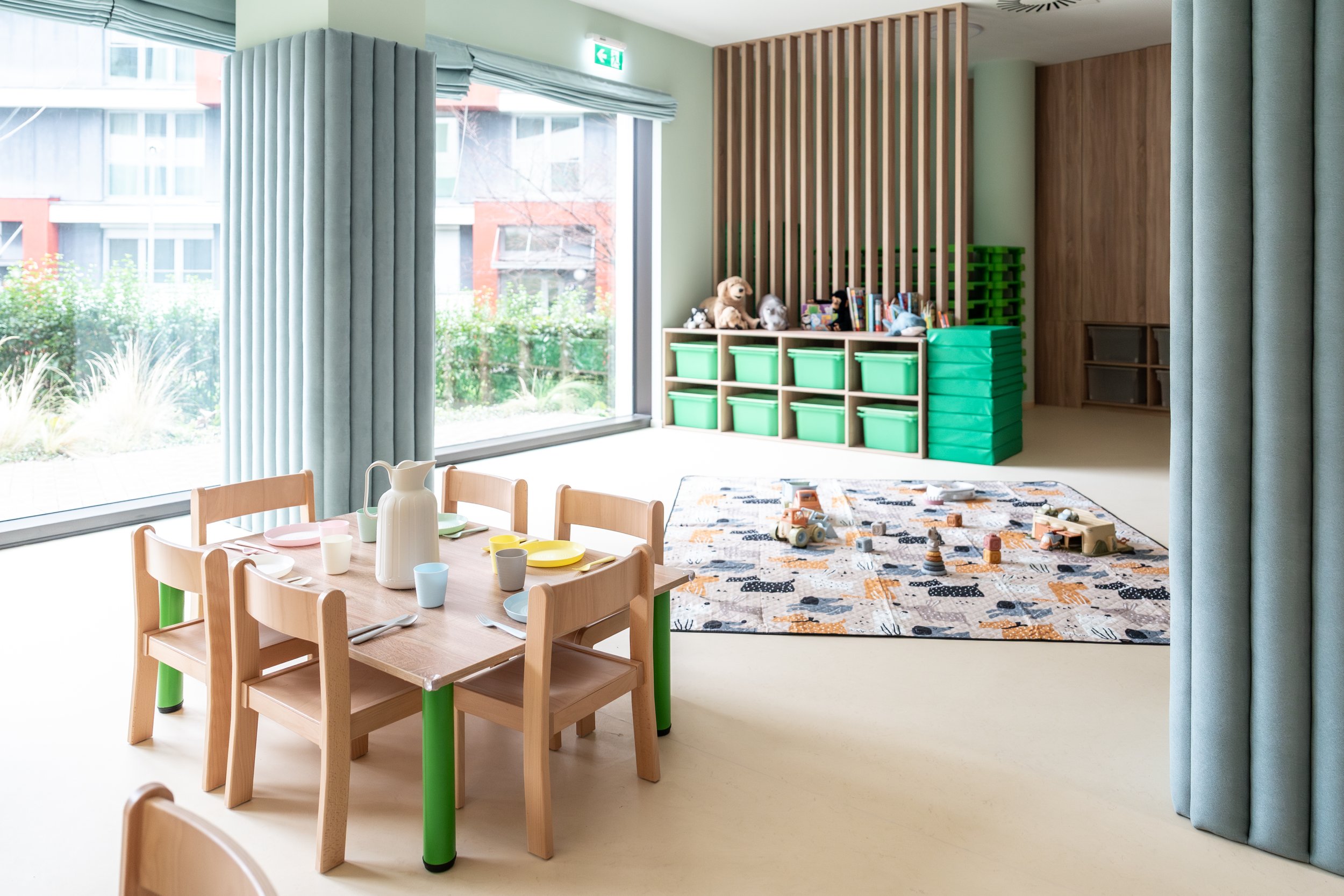 A modern classroom with wooden furniture, large windows with blinds, blue cushions on the benches, and storage bins. The room has ceiling lights and appears bright and inviting.