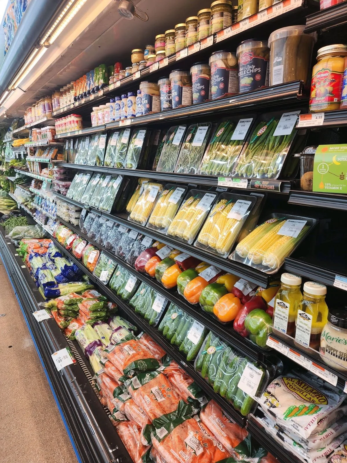 Refrigerated produce section in a grocery store with packaged vegetables including corn, bell peppers, zucchini, and carrots, alongside jars of pickles and canned goods above.