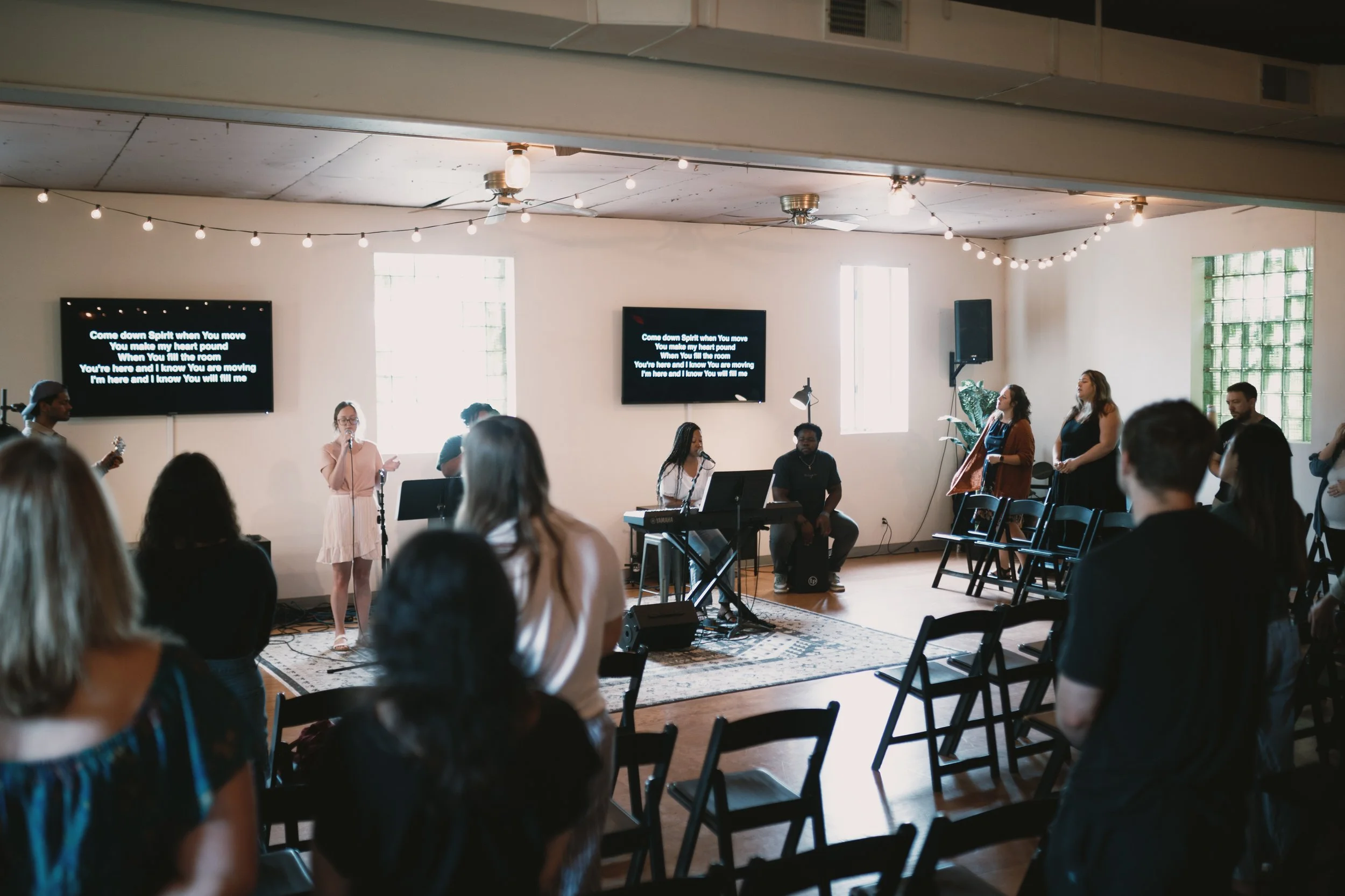 Indoor music event in a room with people gathered, a stage with musicians, and song lyrics displayed on screens.