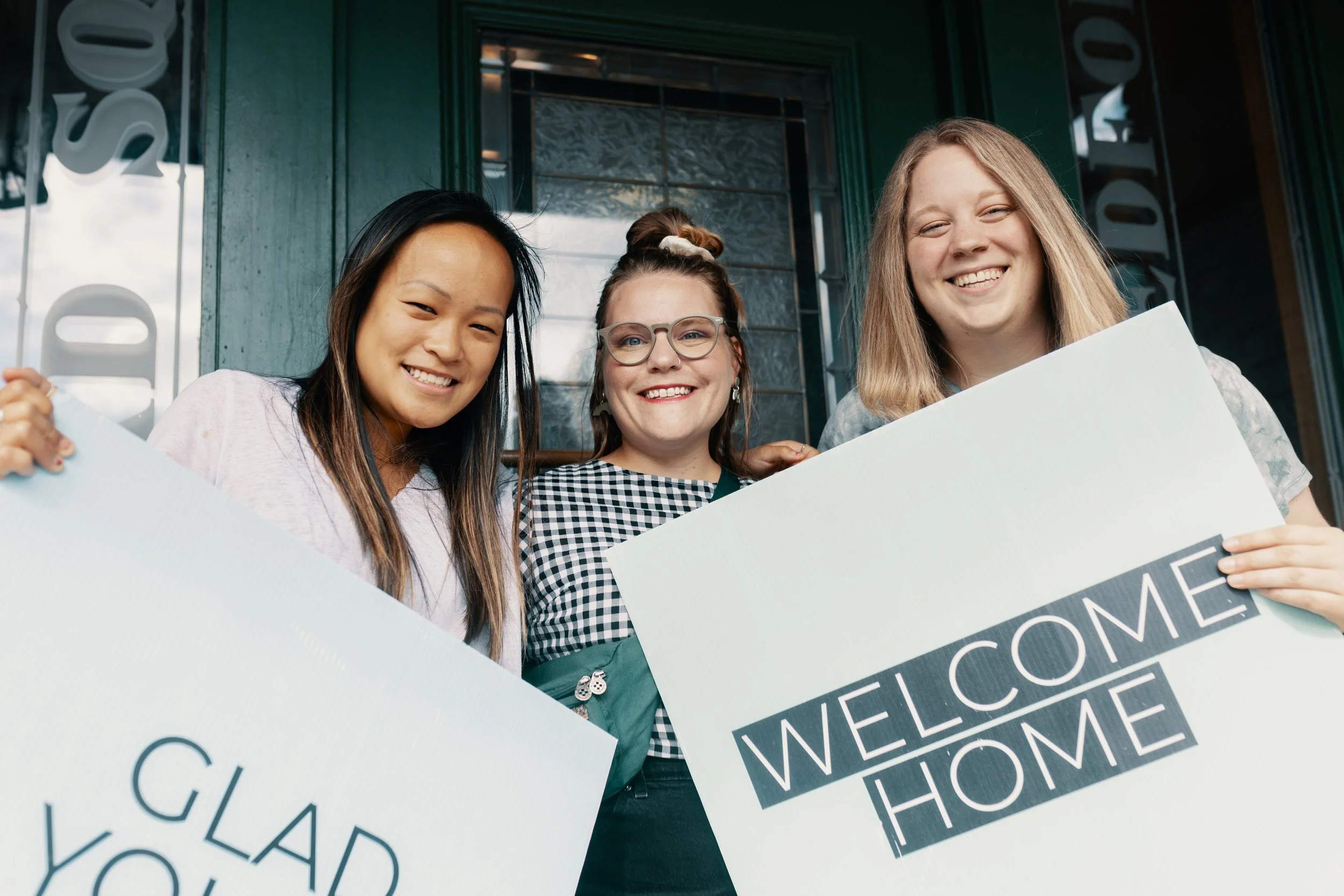 Three people smiling, holding signs that say 'GLAD YOU'RE HERE' and 'WELCOME HOME' in front of a green door.