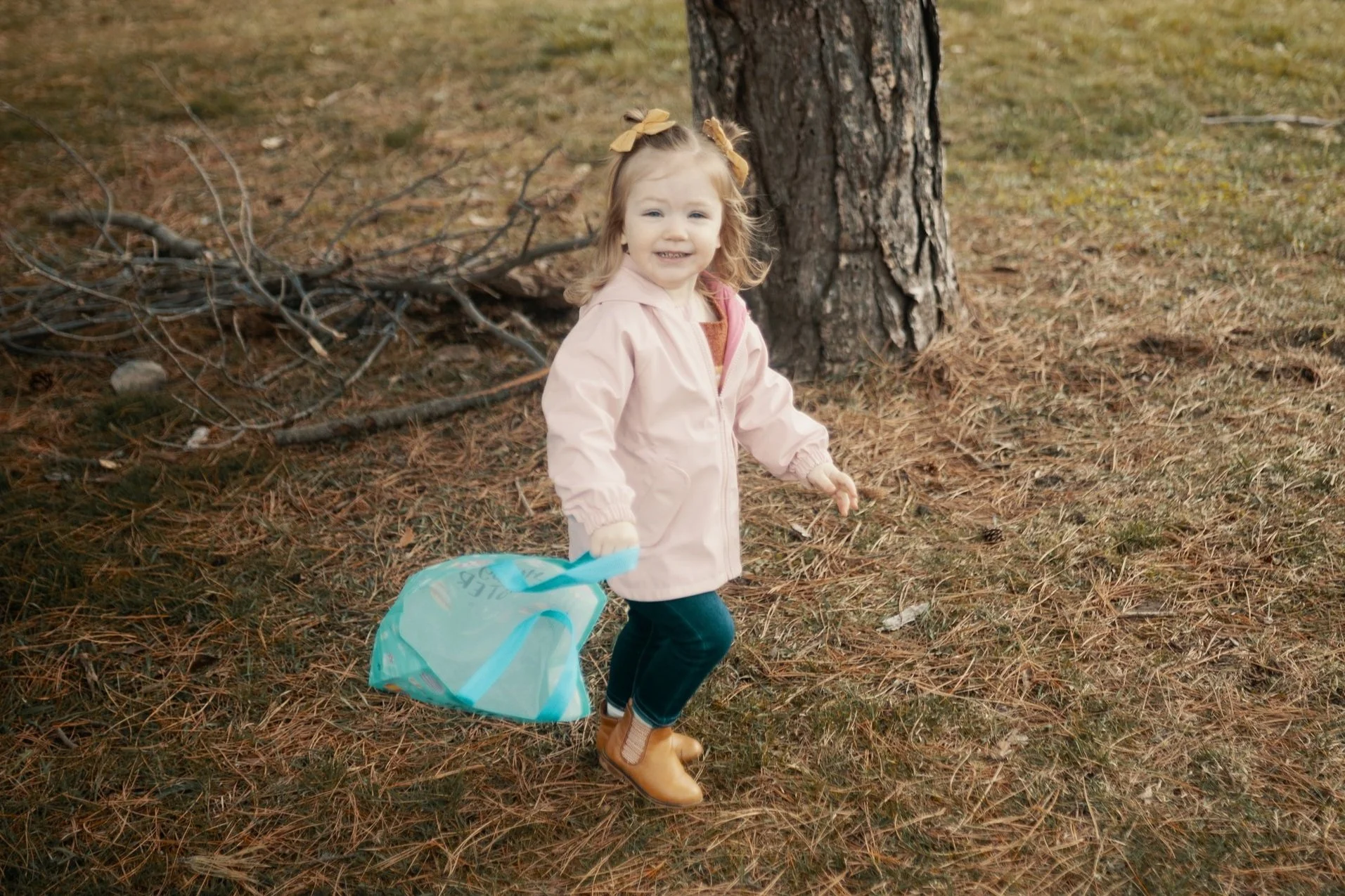 Toddler with a pink jacket and yellow boots carrying a tote bag near a tree in a grassy area.