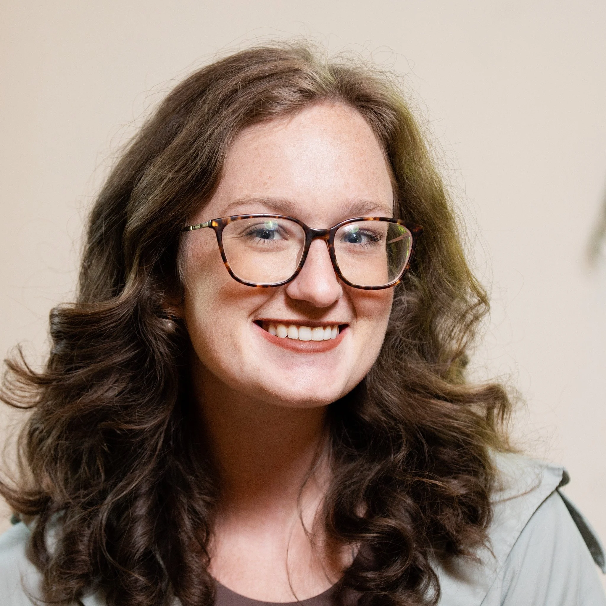 A woman with long, curly brown hair, wearing glasses with tortoiseshell frames, smiling.