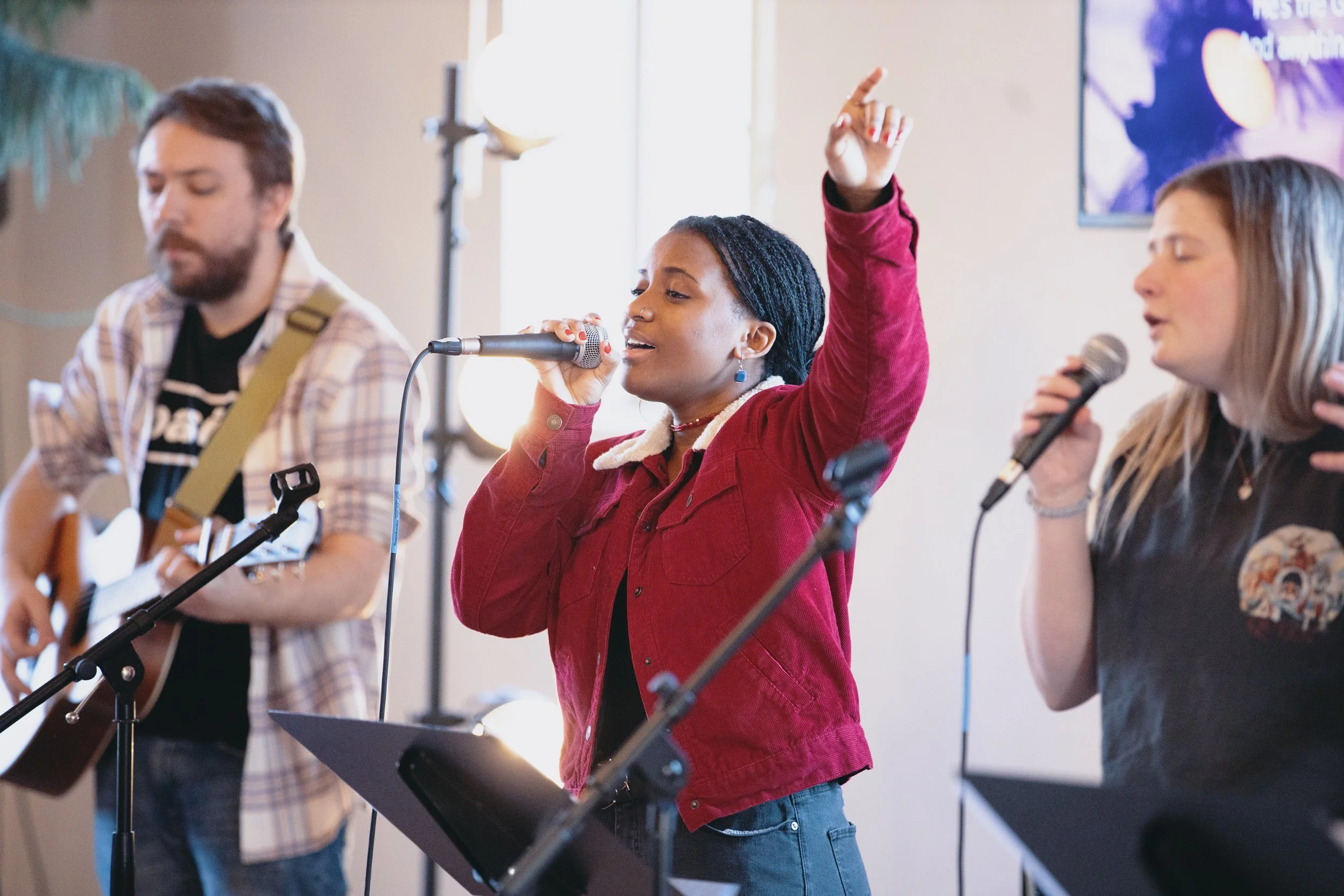 Three musicians performing indoors, featuring a guitarist and two vocalists with microphones. The central vocalist is wearing a red jacket and raising one arm.