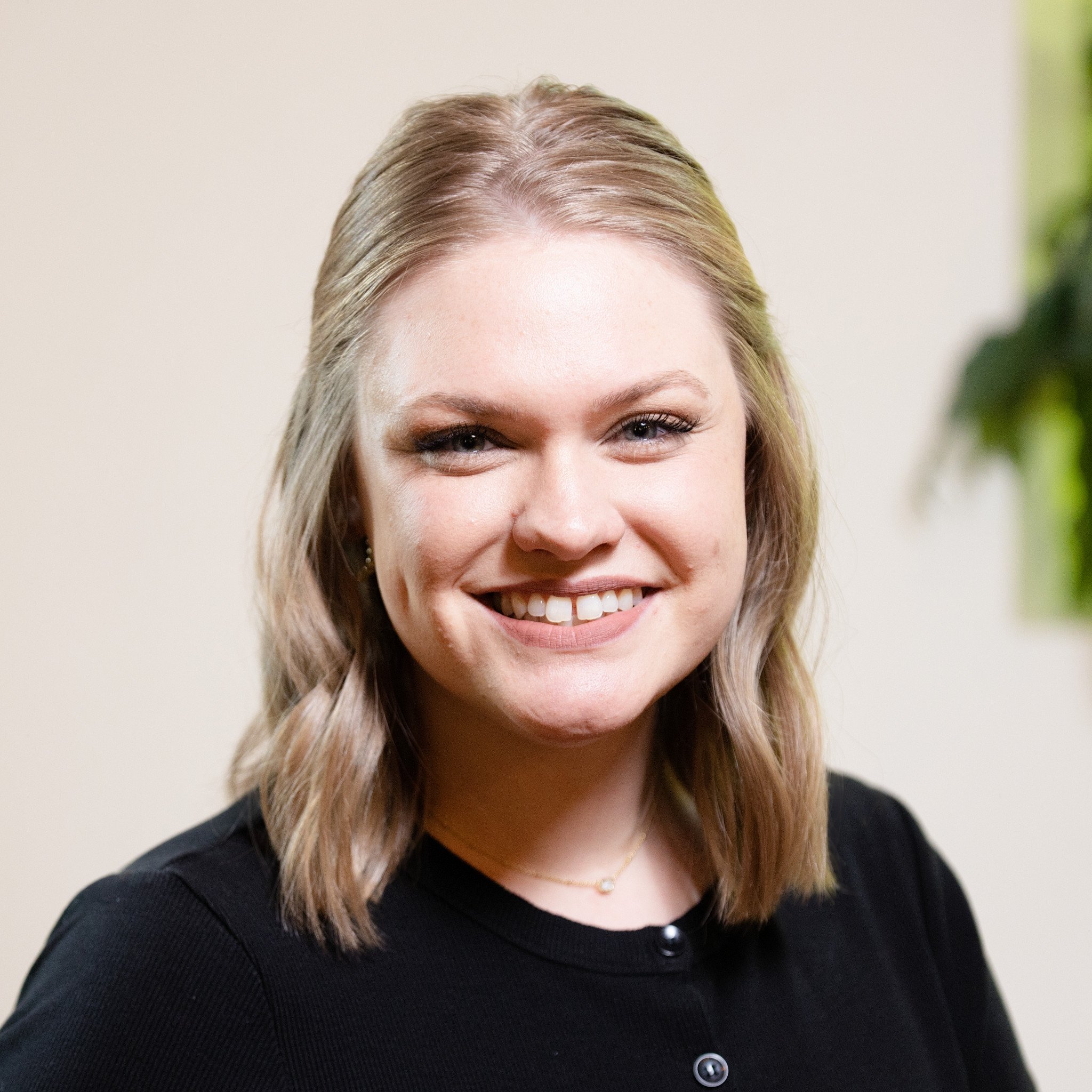 A smiling woman with shoulder-length blonde hair wearing a black top, gold necklace, and earrings, standing indoors with a blurred background and some greenery.