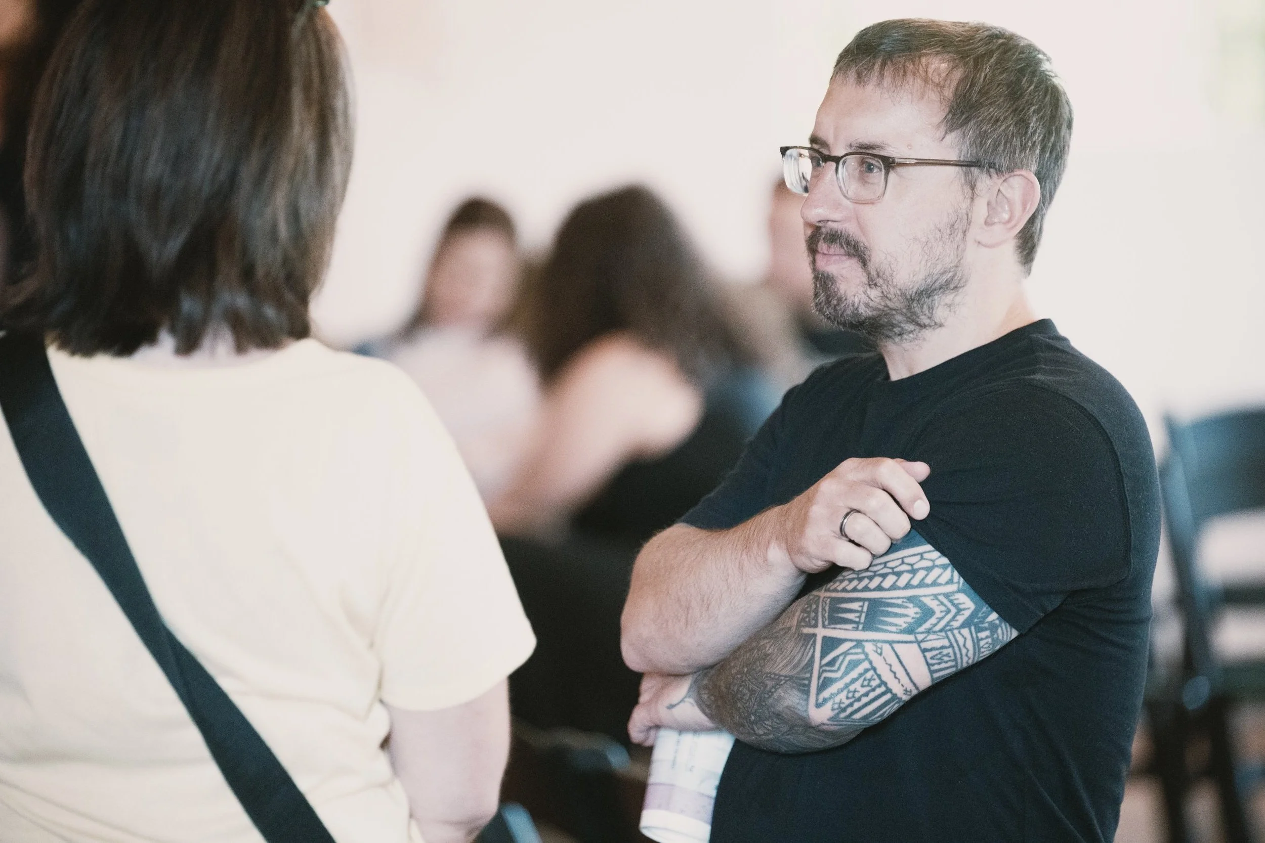 Two people having a conversation; one with a tattooed arm and wearing glasses, the other with a yellow shirt and back facing the camera.