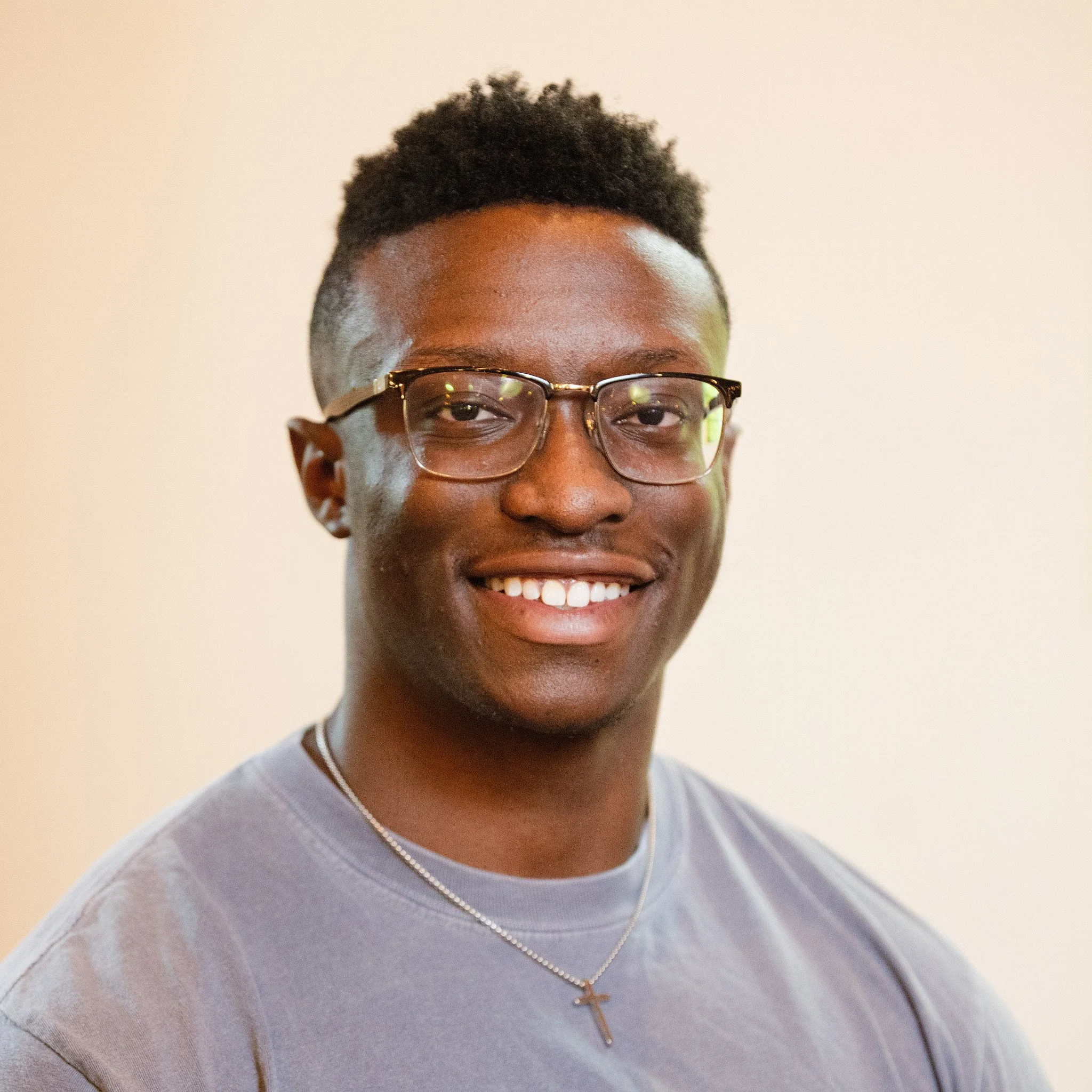 A young man with glasses smiling against a plain, light-colored background.