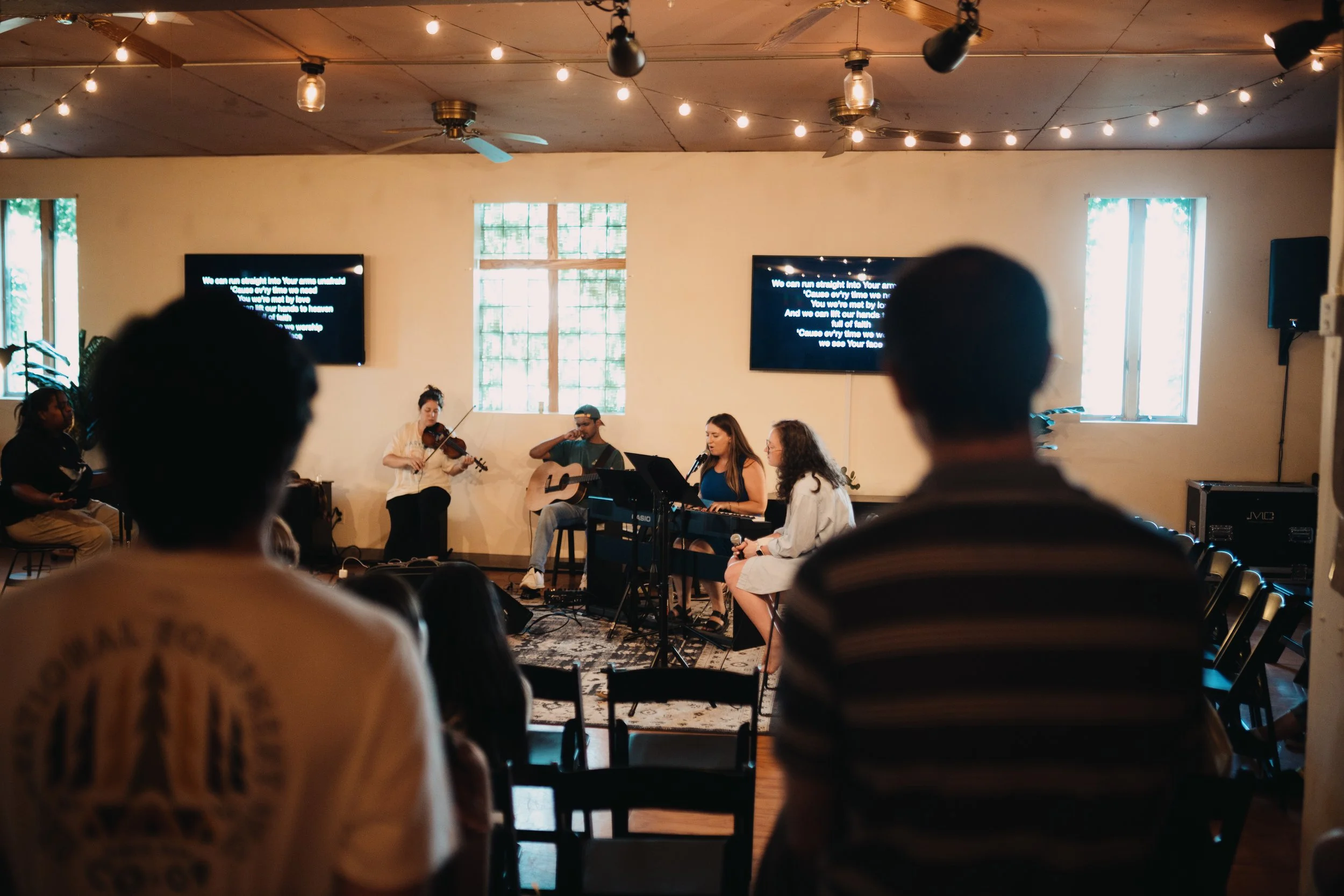 A group of musicians performing in a cozy indoor setting with string lights, a ceiling fan, and two TV screens displaying lyrics, while an audience watches.