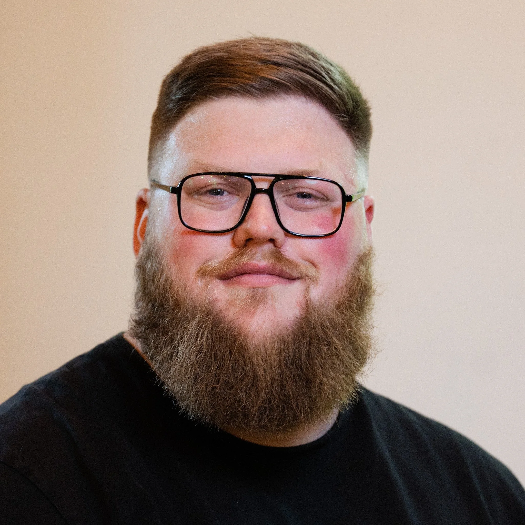 Man with a beard and glasses wearing a black shirt, sitting indoors next to a large potted plant.