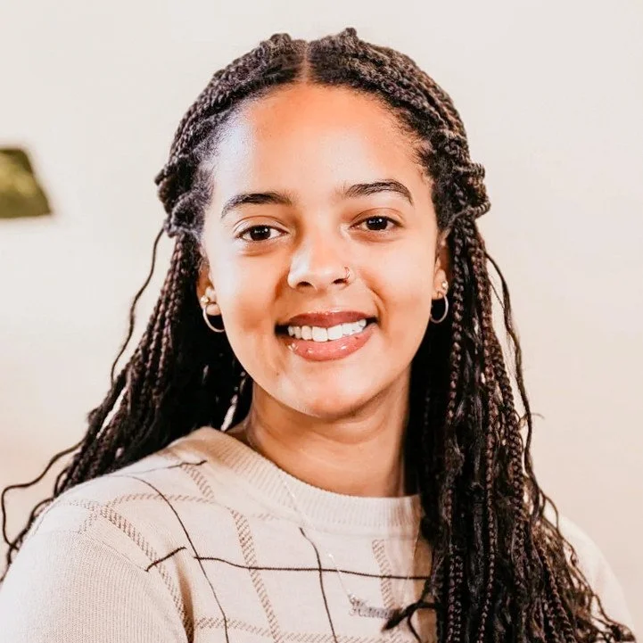 Smiling woman with braided hair wearing a beige checkered sweater, indoor background with a plant.