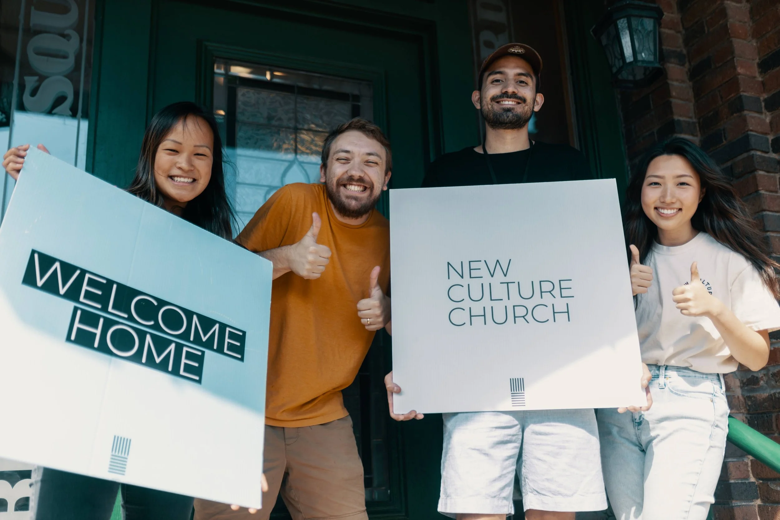 Group of four people smiling, holding signs reading "Welcome Home" and "New Culture Church" outside a building.