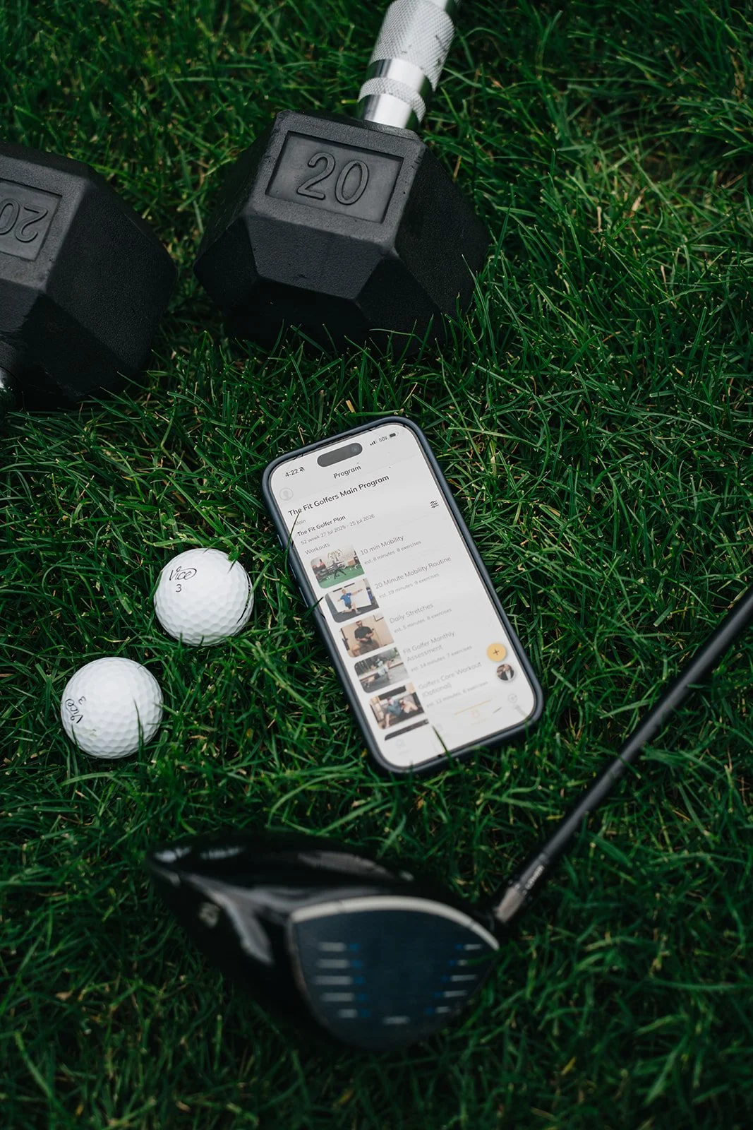 Golf equipment including two black dumbbells labeled 20, two white golf balls marked 3 and 4, a golf club, and a smartphone on grass displaying a golf workout program.