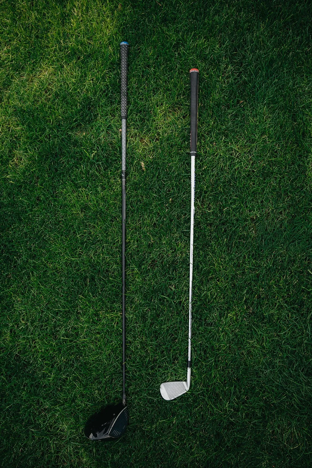Two golf clubs lying on green grass, one with a black head and the other with a silver head.