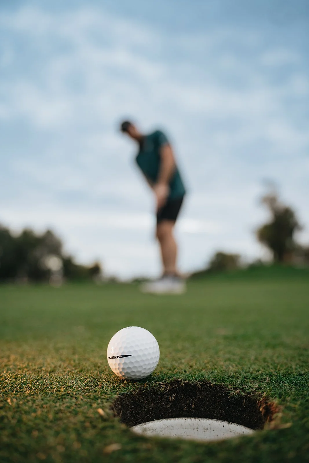 Close-up of a golf ball on the edge of a hole on a golf green, with a golfer in the background preparing to take a shot.