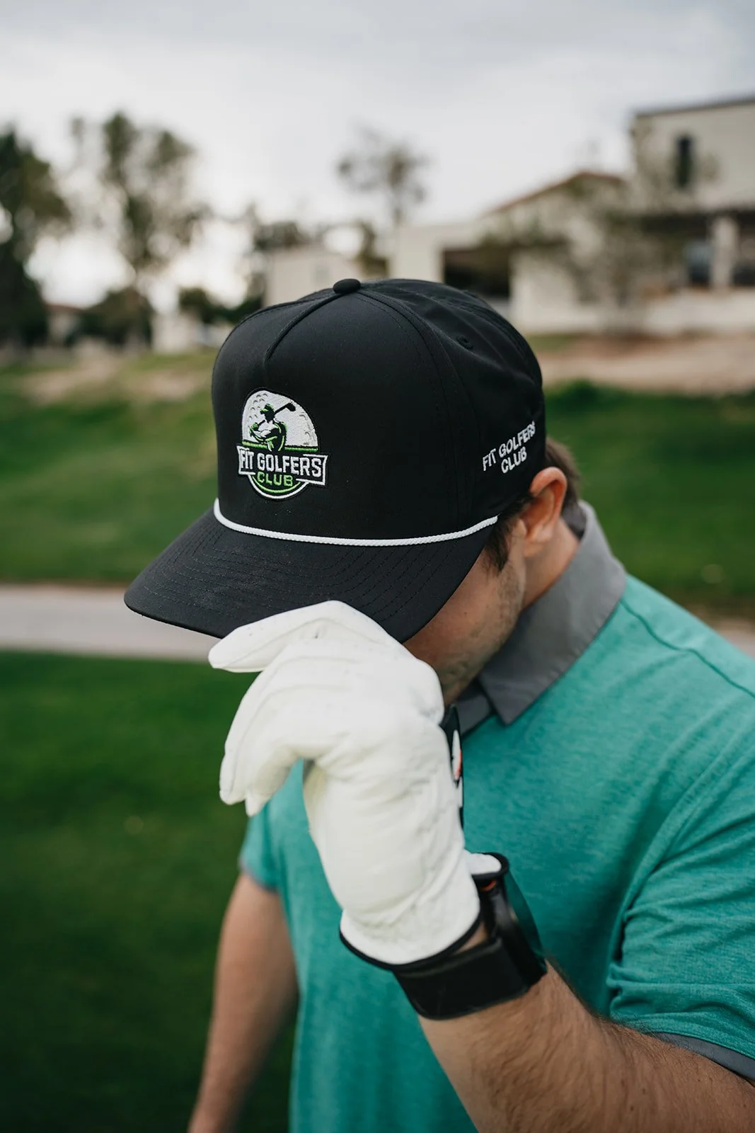 Close-up of a man wearing a black golf cap with 'Fit Golfers Club' logo, a teal shirt, white glove, and black watch, on a golf course with blurred houses in the background.
