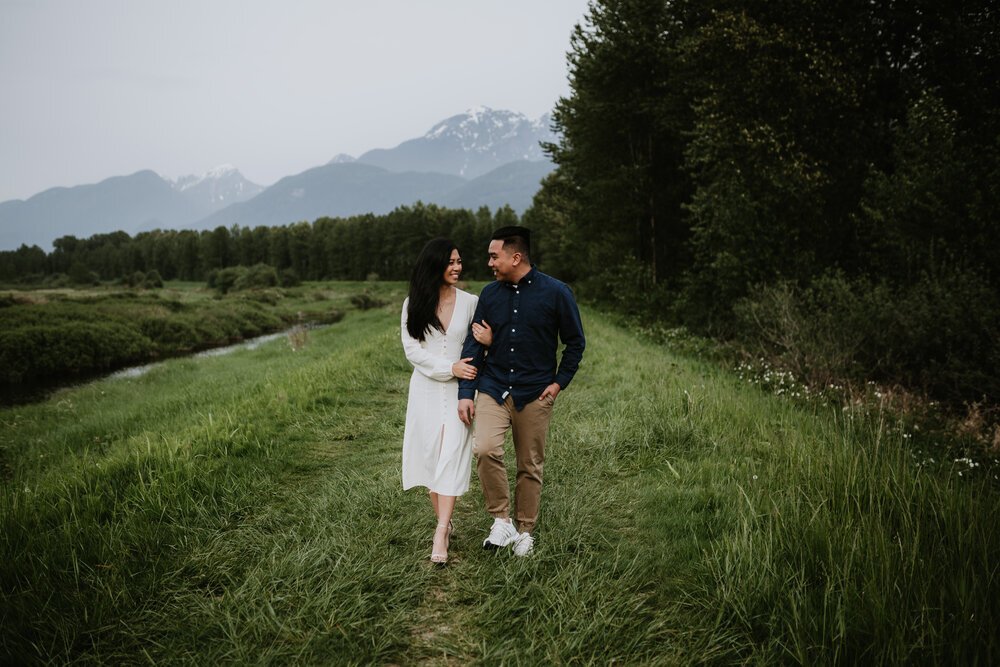 Engaged couple walk sided by side while gazing into each other's eyes at Pitt Lake in Pitt Meadows.