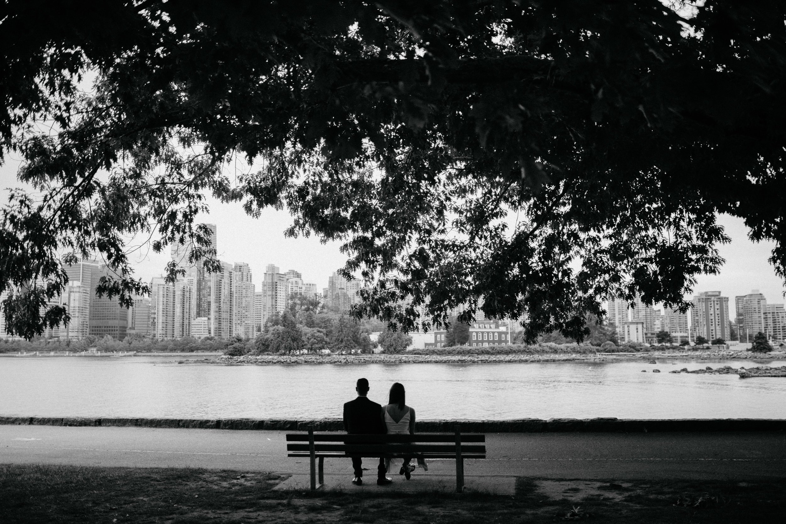 Engaged couple sitting on a bench by the seawall at Stanley Park, while looking at the view of coal harbour.