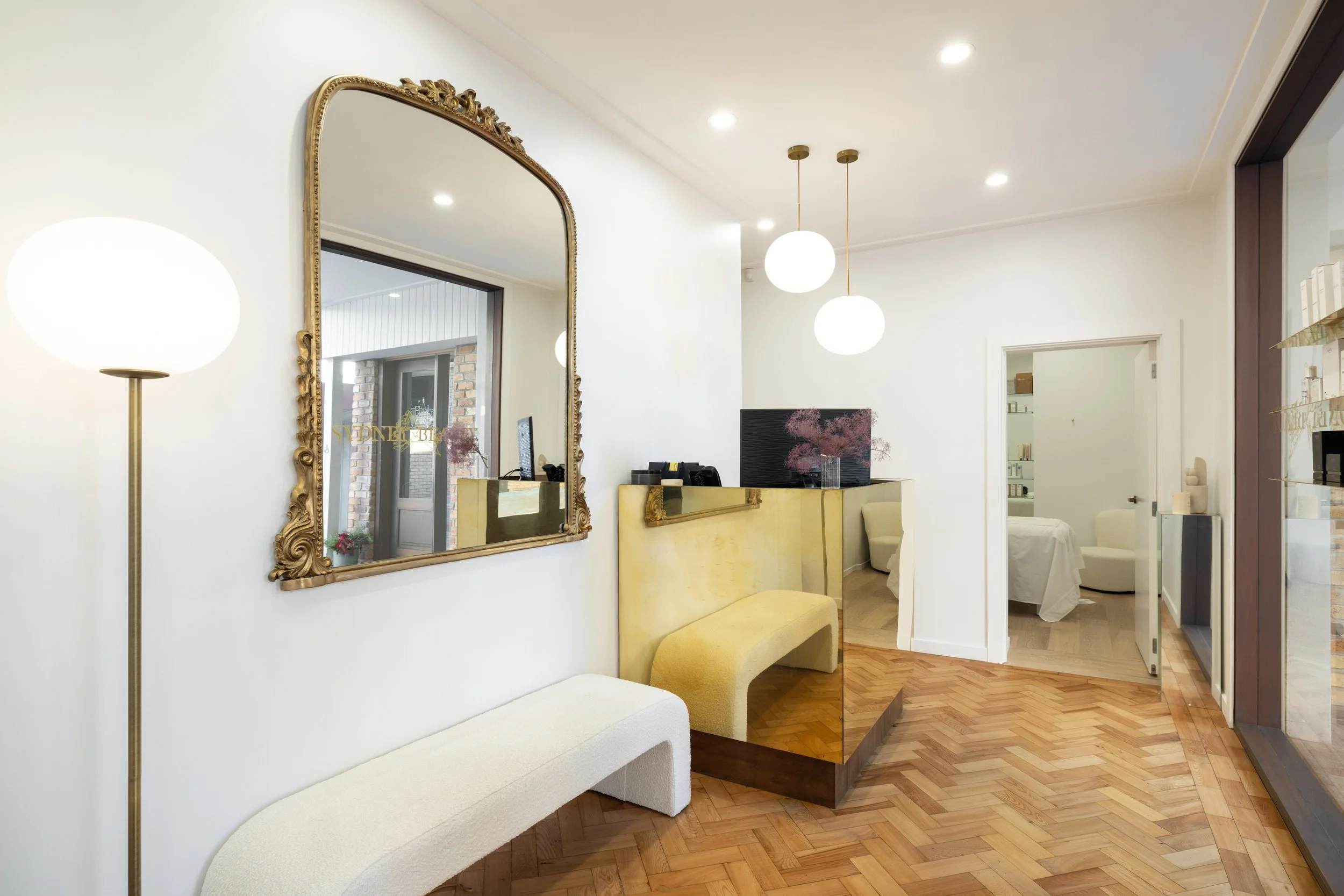 Interior of a modern, minimalist waiting area with a large decorative mirror, white walls, wooden floor, and seating. There are hanging spherical light fixtures and a gold-colored reception desk in the background.