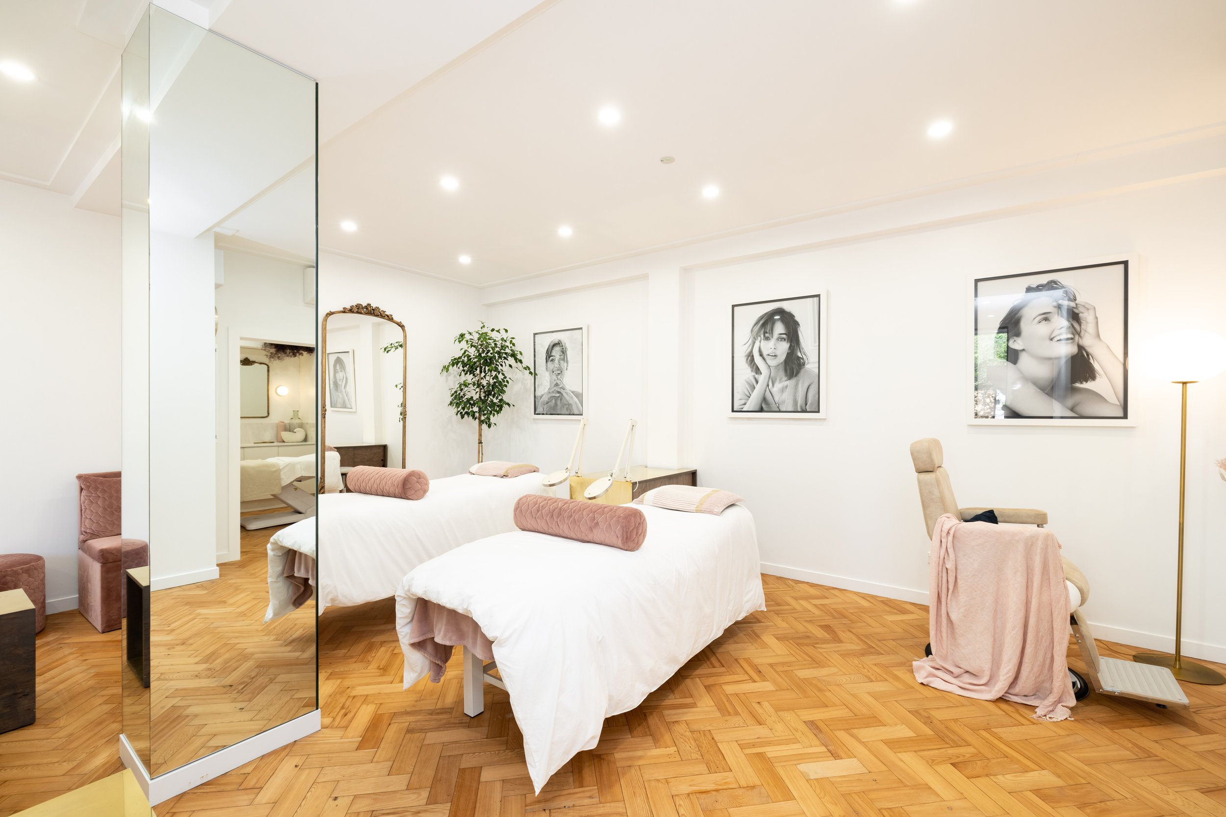 A bright, modern bedroom with white walls and hardwood flooring. It features two twin beds with white bedding, pink bolsters and pillows. Framed black and white portraits hang on the wall. There is a large mirror, a potted plant, a beige armchair wit