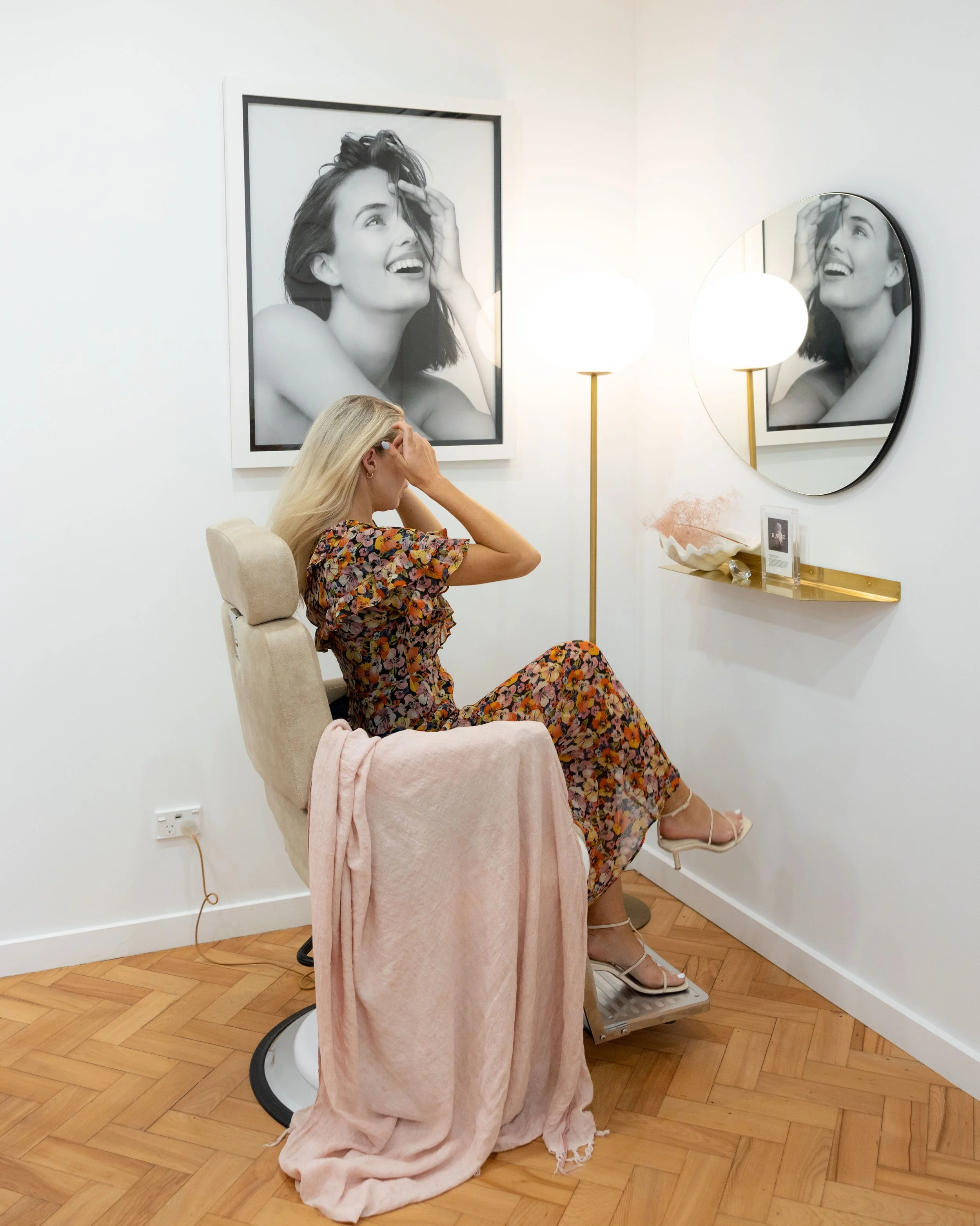 A woman with long blonde hair sitting in a salon chair, covering her face with her hand, in front of a mirror, with black and white portraits of a smiling woman with hand in hair on the wall behind her, in a modern salon with wooden flooring and soft