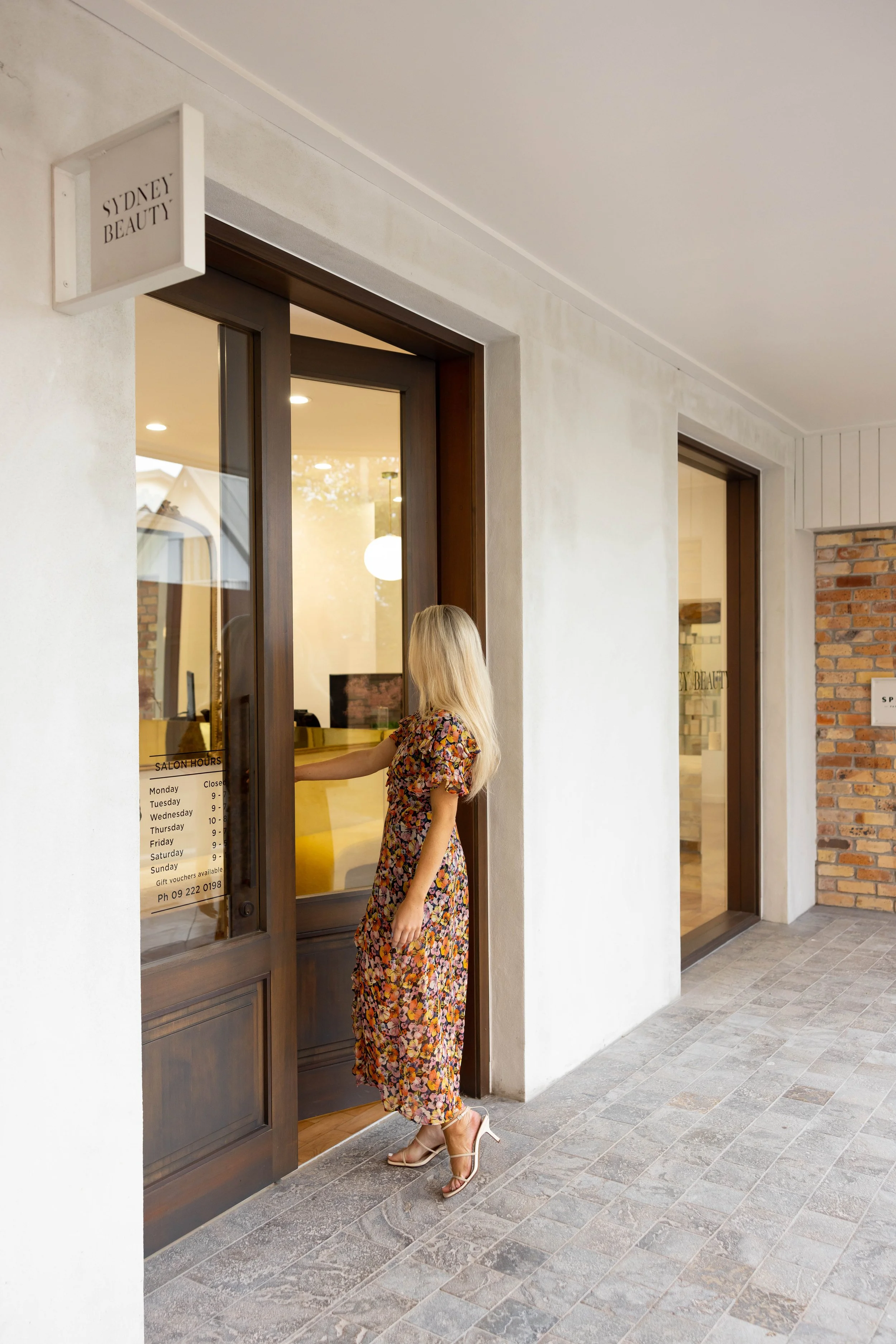 A woman in a floral dress and high heels is opening the door to a beauty salon named Sydney Beauty, which has a sign detailing its hours of operation.