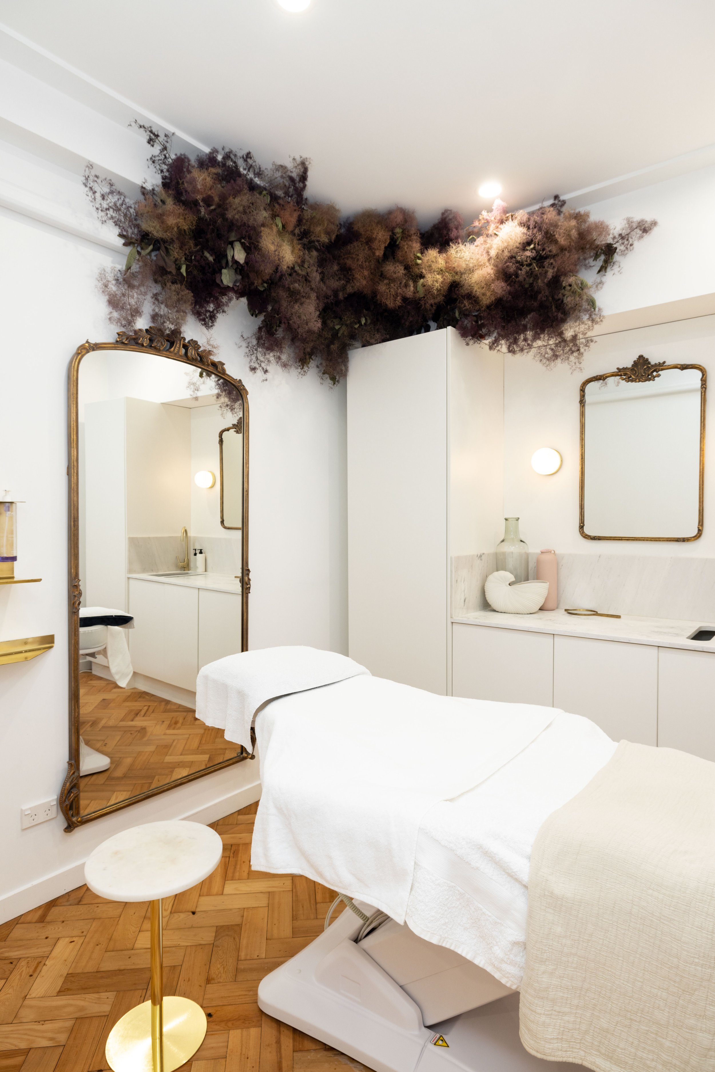 A spa or treatment room with a massage table draped in white linens, a large ornate mirror, and decorative dried flowers on the ceiling, with wooden flooring and minimalist decor.