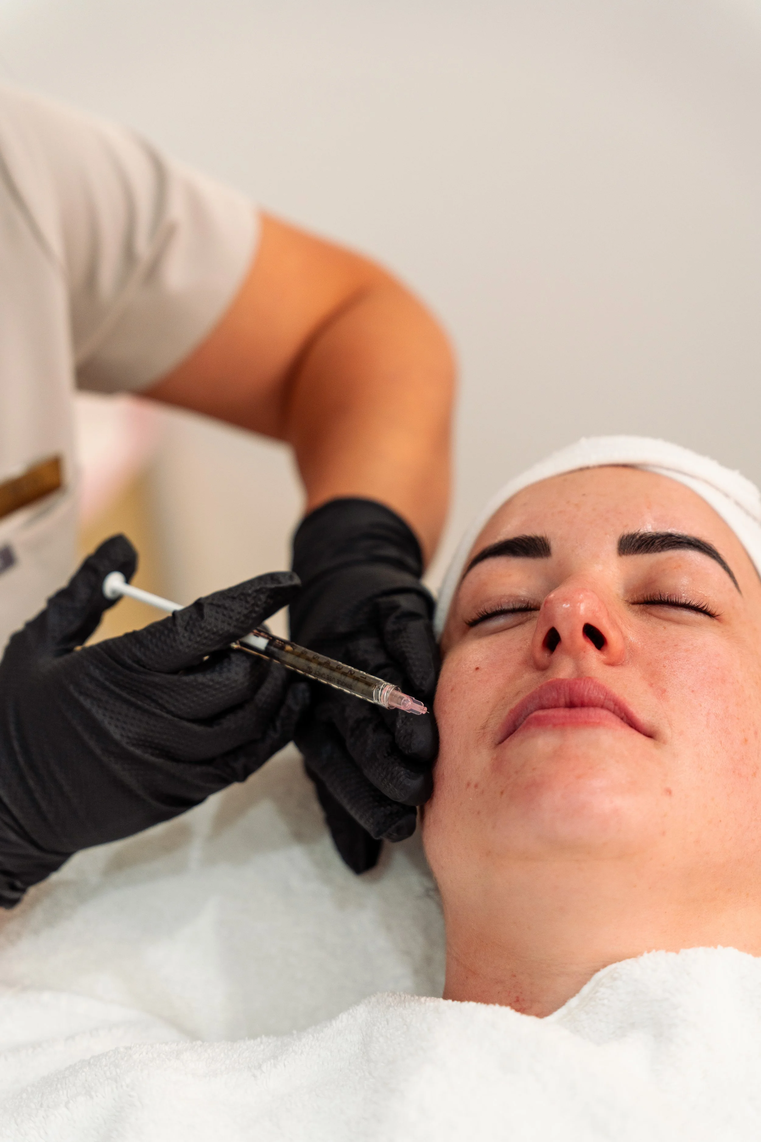 A woman receiving a facial injection, lying down with her eyes closed, while a medical professional administers the shot using a syringe.