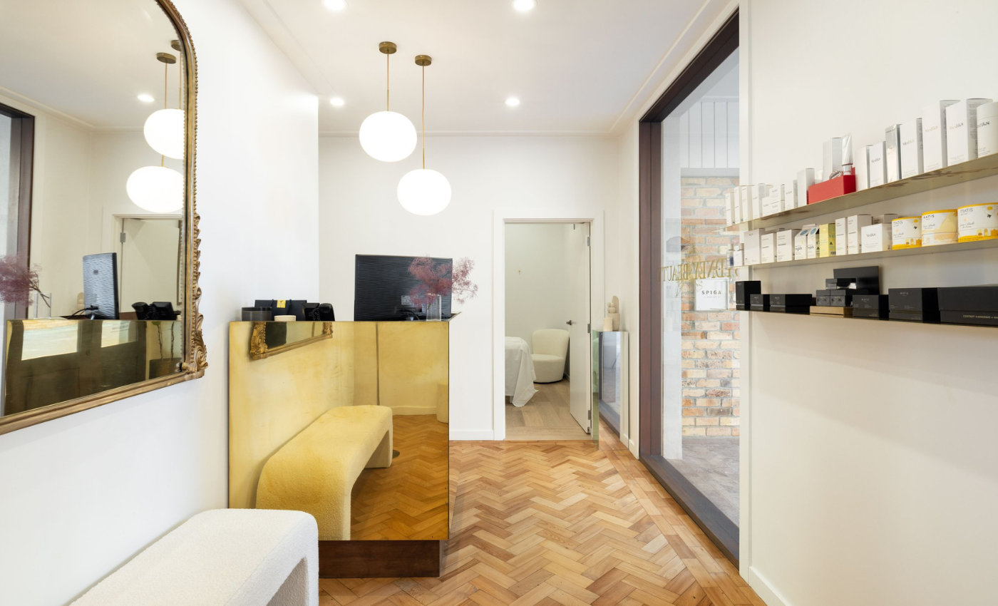 A modern reception area with a mirrored gold-front counter, yellow velvet seating, white walls, a large mirror, white spherical pendant lights, and a shelf with boxes and black items, leading to a small room with white chairs.