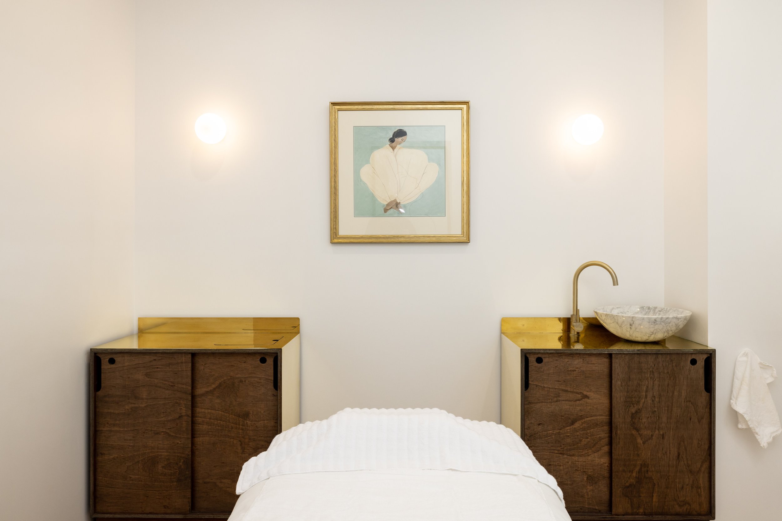 A minimalist bedroom with white walls, a framed artwork of a woman in white sitting with her back turned, two wall-mounted lights, two wooden cabinets on either side of a bed, and a stone basin sink with a brass faucet on the right cabinet.